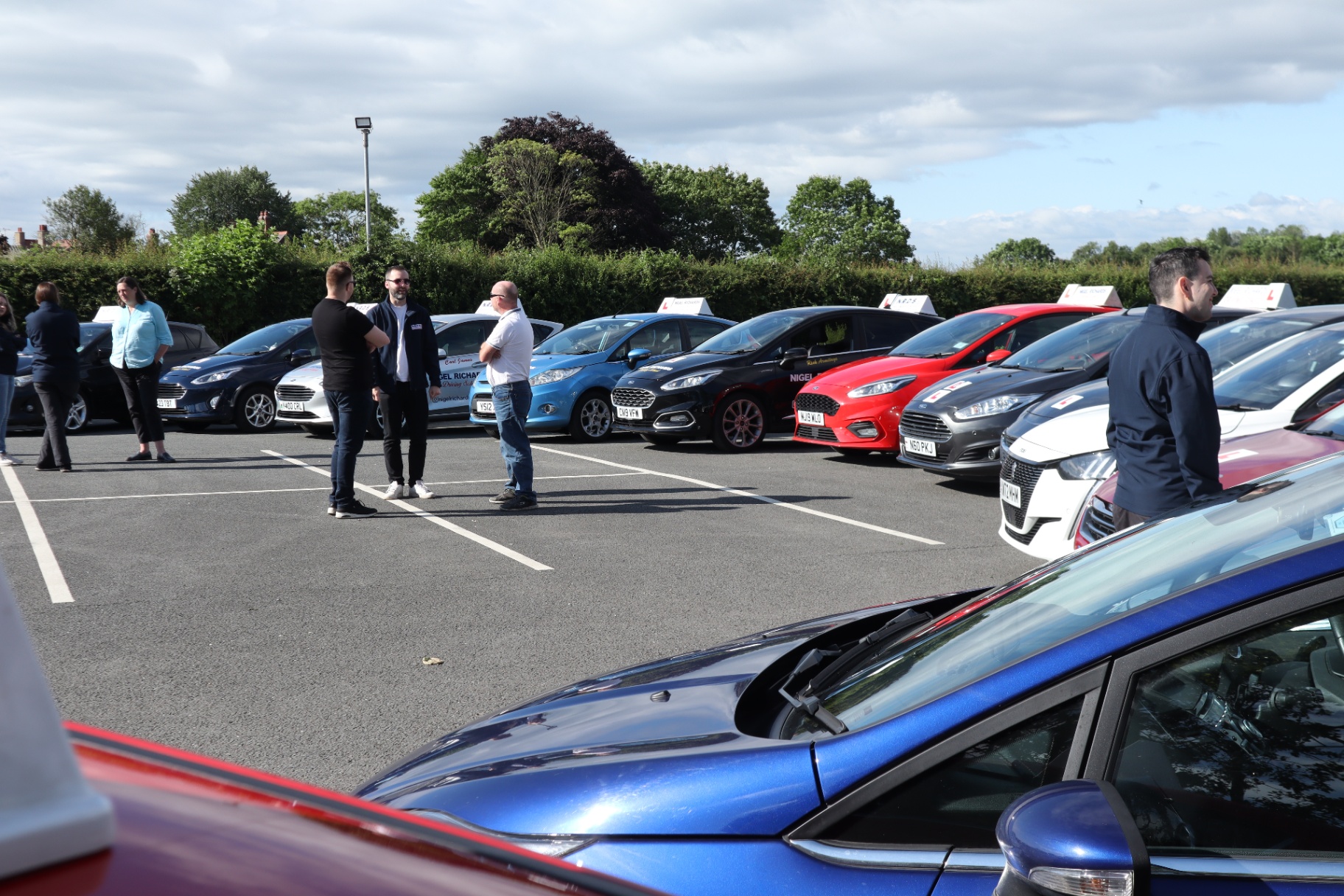 Nigel Richards Driving School Instructors Gathering Several Nigel Richards Driving School instructors and staff are gathered in a paved car park, conversing beside a line of dual-control cars all branded with L plates and driving school roof signs. The vehicles represent a modern, well-maintained fleet for both manual and automatic tuition.