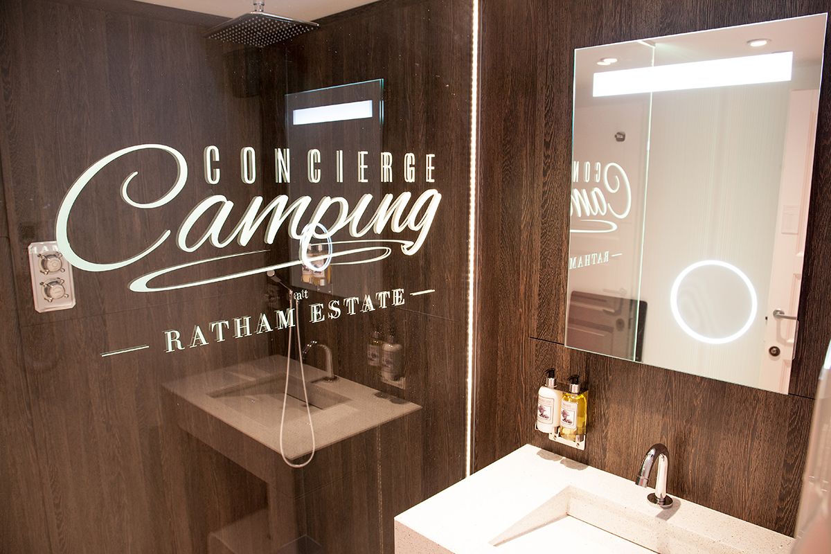 Modern campsite bathroom at Concierge Camping, Ratham Estate. Features include stylish dark wood walls, illuminated mirror, rain shower, and branded luxury toiletries. Highlights the premium amenities available for caravaners looking for high-quality campsite facilities.
