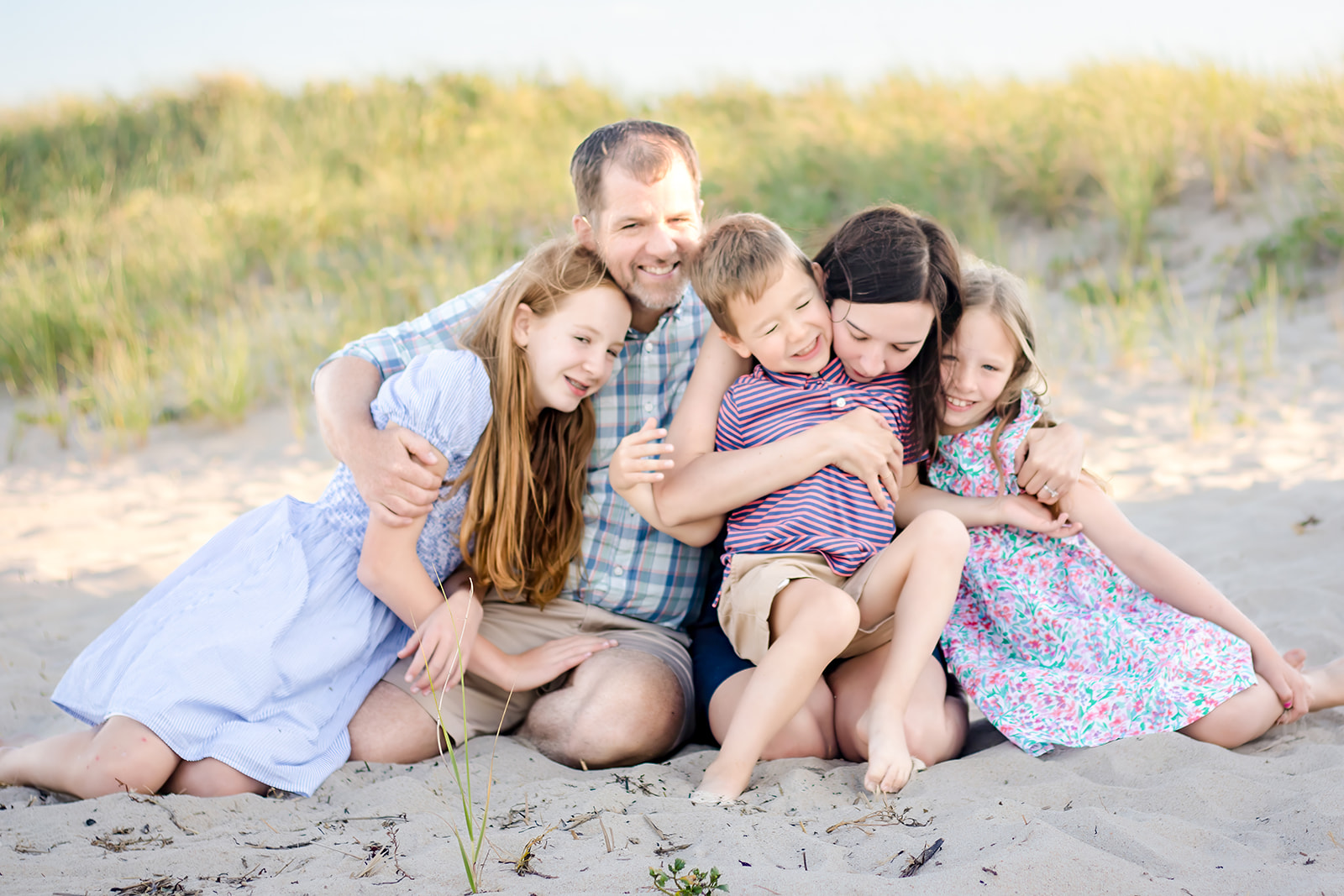 A family of five sits together on a sandy Cape Cod beach, surrounded by tall grass, sharing a group hug.