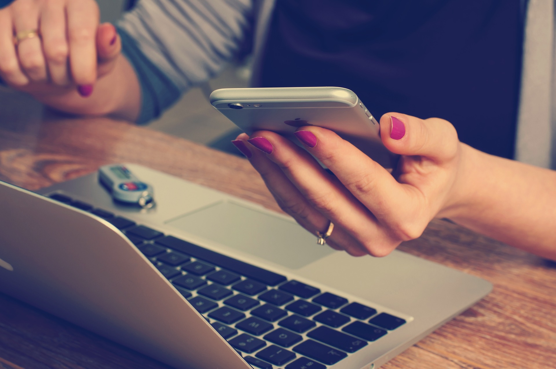 A person with painted nails works at a wooden desk with a laptop, a USB drive, and a smartphone in hand