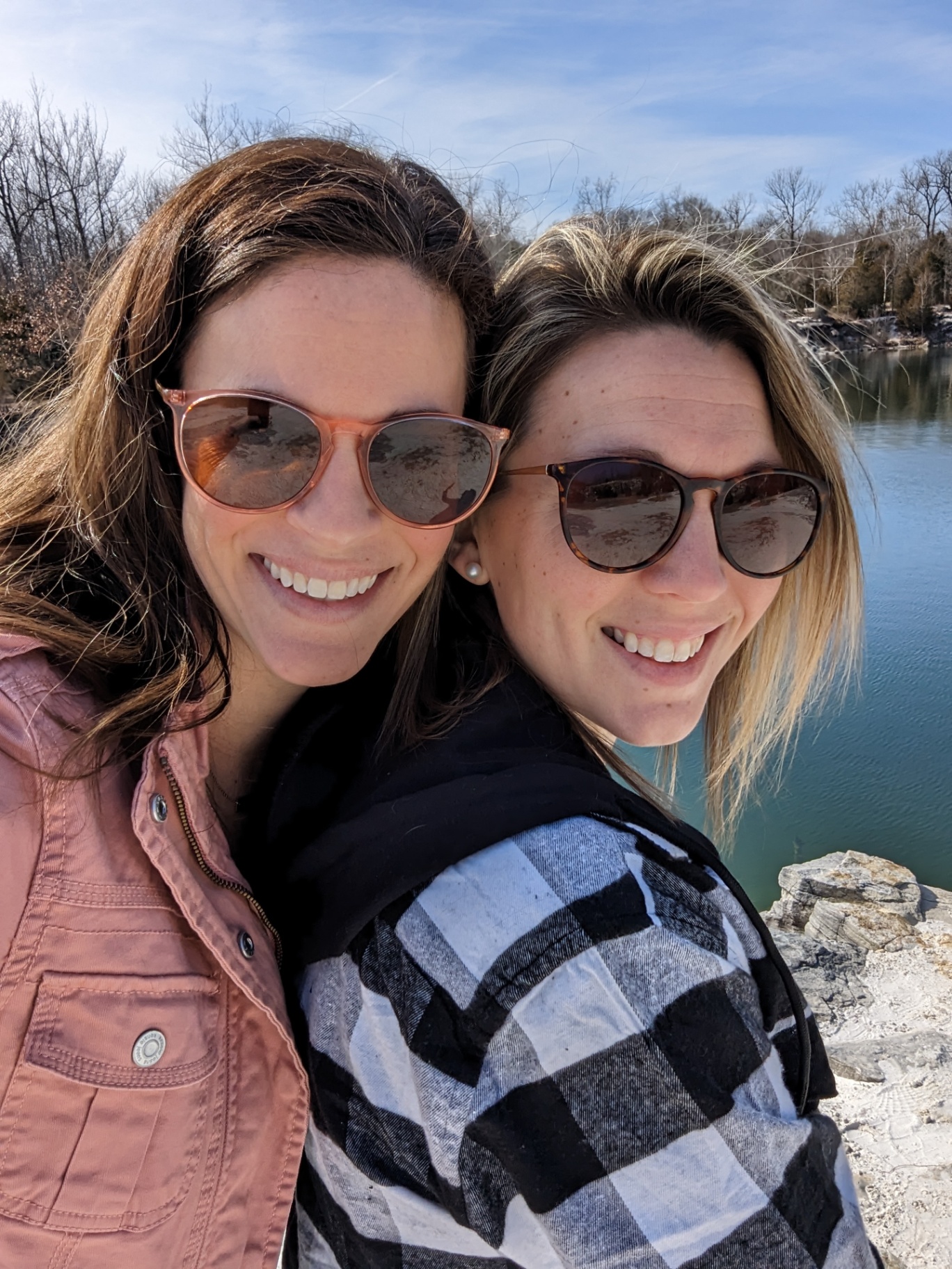 Two women wearing sunglasses and casual jackets smile while standing close together outdoors by a lake