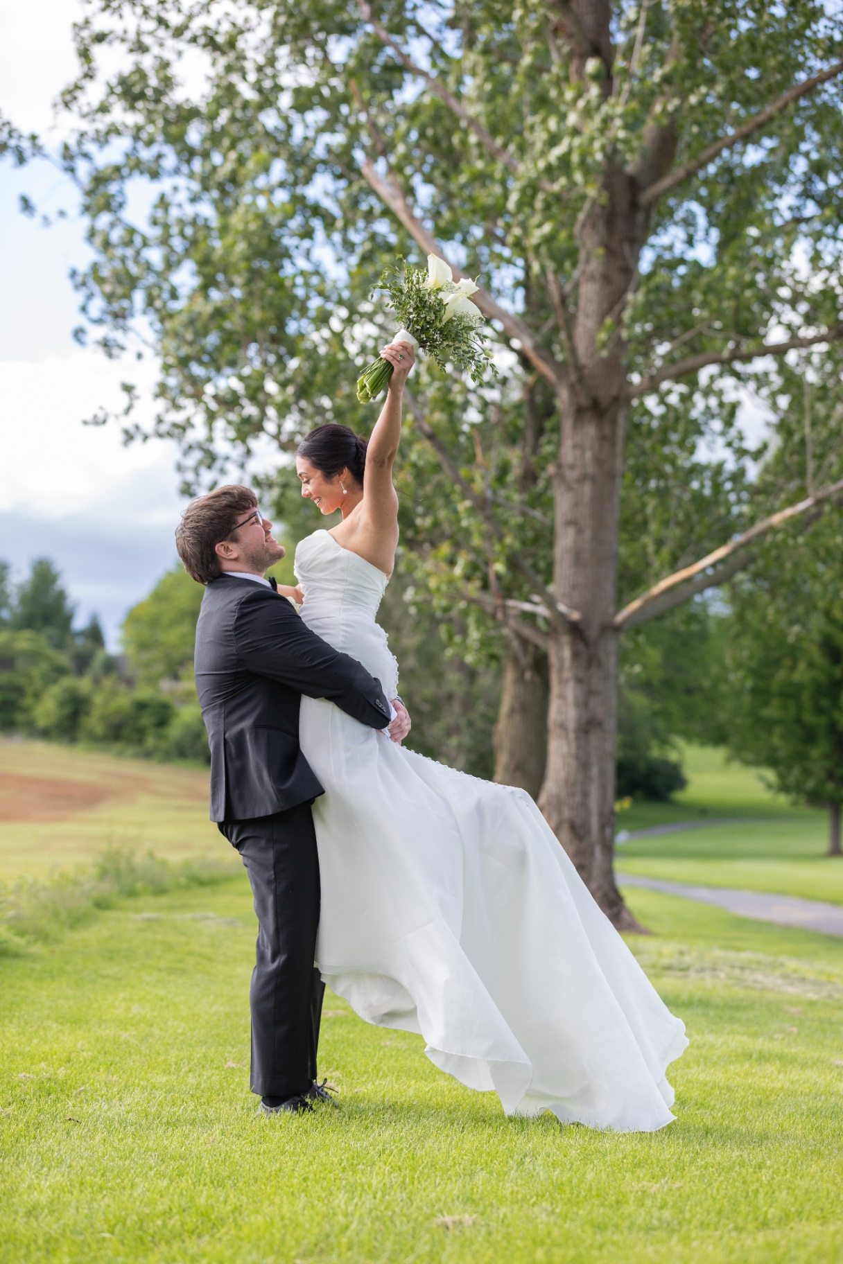 A groom joyfully lifts his bride outdoors surrounded by lush greenery