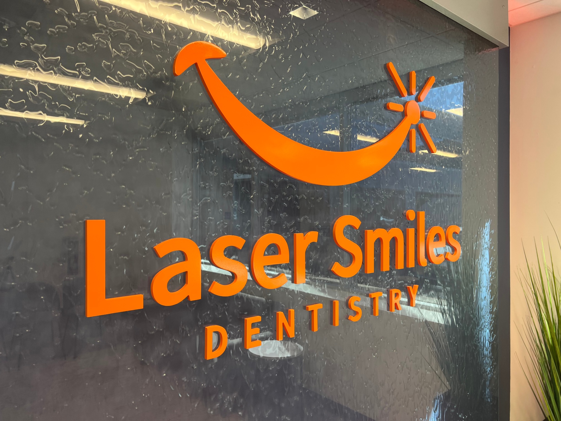 Custom Waterwall at Laser Smiles Dentistry A custom waterwall installed at a dental practice, featuring a backlit 'Laser Smiles Dentistry' logo on the glass surface. The waterwall uses exclusive L2O Liquid running down in a clear, maintenance-free sheet, creating a sophisticated, modern entrance and highlighting the company's branding.
