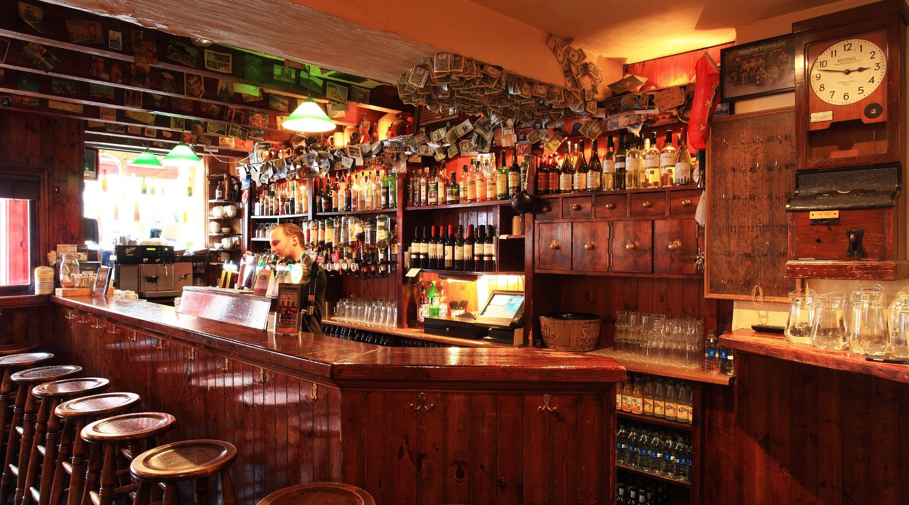 Traditional Irish Pub Interior - Kenmare Traditional Irish pub interior in Kenmare with polished wooden bar, a row of wooden stools, shelves stocked with spirits, wines, and glassware, and unique decor featuring international currency notes and photos. Welcoming atmosphere showcasing local hospitality for guests staying at Kenmare Rentals, within walking distance of our self-catering holiday homes.