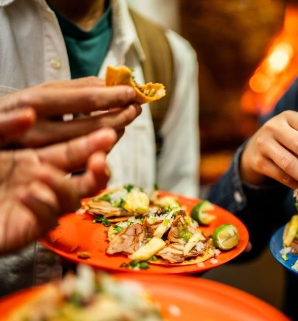 Groups of festival-goers enjoying vibrant global street food, holding tacos garnished with fresh toppings and limes on bright orange and blue plates.
