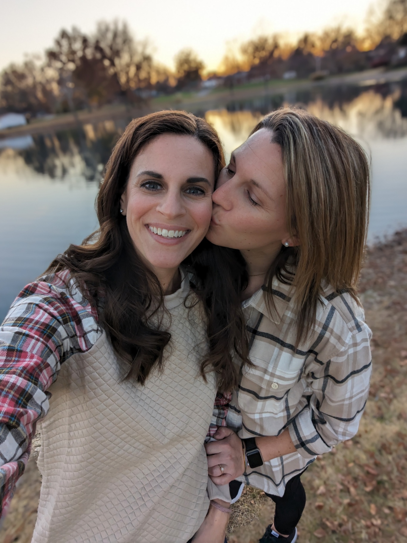Two women stand outdoors by a lake at sunset, one smiling at the camera while the other gently kisses her on the cheek. They are holding hands and wearing casual plaid shirts, conveying warmth, connection, and authenticity.