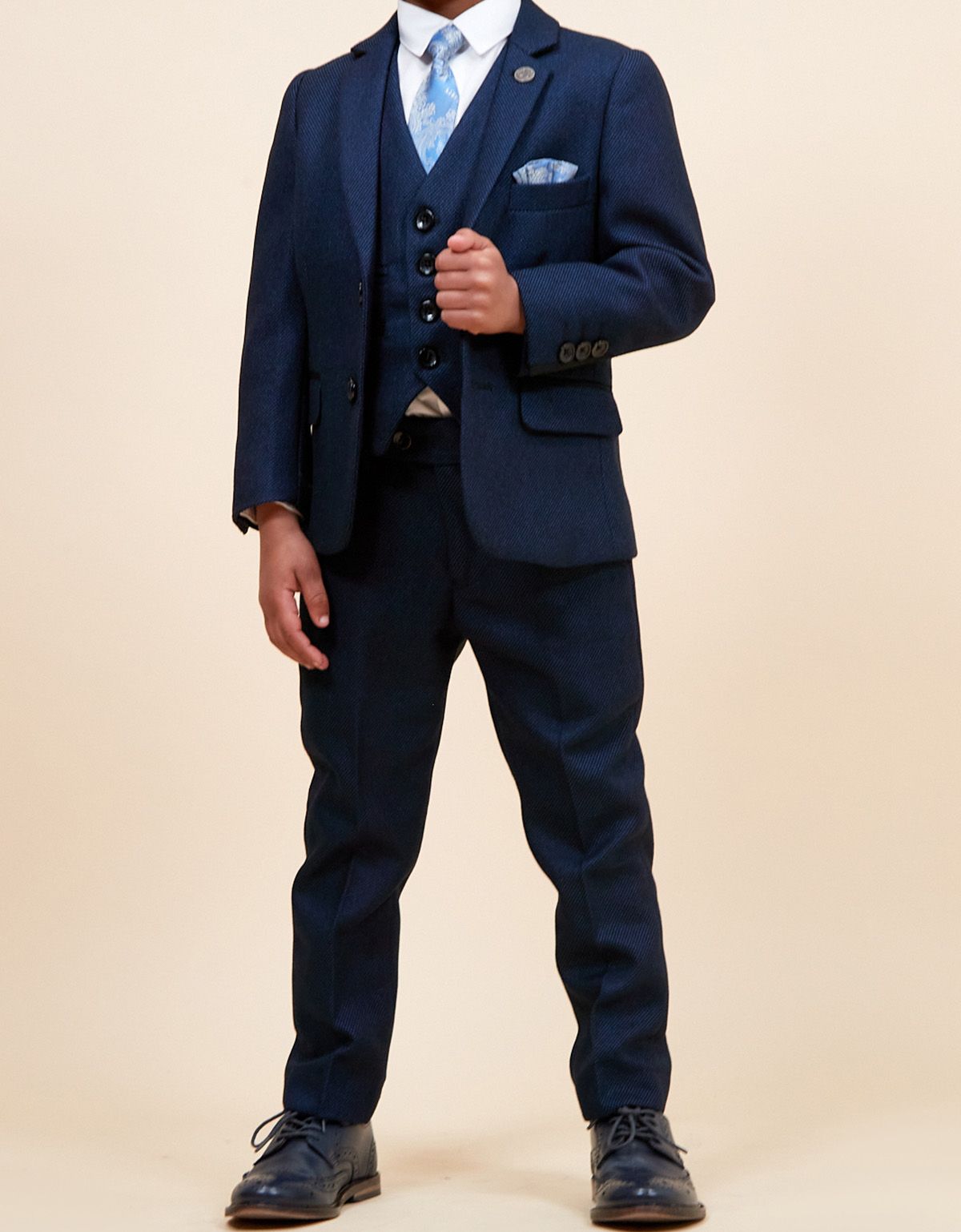 A young boy models a sharp three-piece navy suit with matching waistcoat, white shirt, blue floral tie, and pocket square.