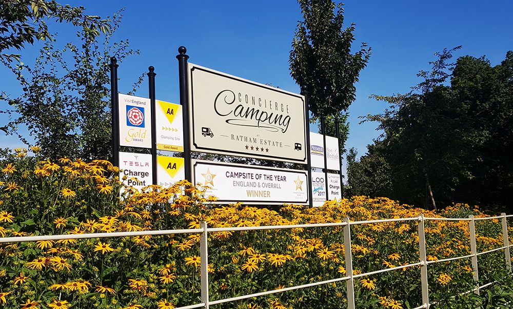 Concierge Camping at Ratham Estate entrance sign surrounded by yellow flowers, showcasing awards such as AA Gold, Visit England Gold Award, and 'Campsite of the Year, England & Overall Winner.'