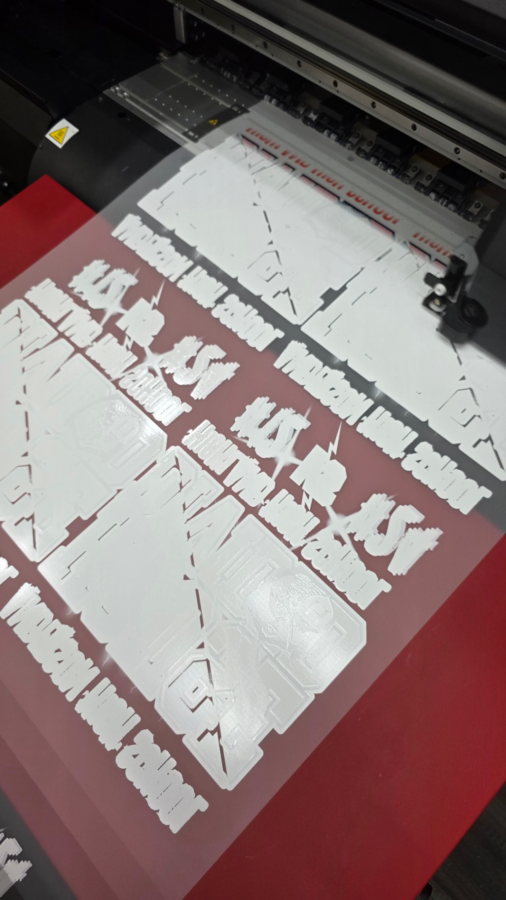 DTF Gang Sheet Printing A DTF printer produces a gang sheet loaded with custom white toner ink transfers. Multiple graphics and text elements are efficiently arranged on a single clear film, ready for cutting and pressing onto apparel. This setup demonstrates the high-quality, crisp results and versatile output of Press Ready DTF's gang sheet printing service.