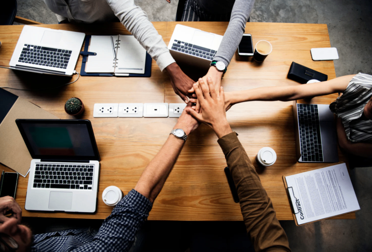 Arc & Beat Lab A diverse group of filmmakers collaborates around a table with laptops, notebooks, coffee cups, a contract, and power outlets, joining hands in a gesture of teamwork and mutual support, reflecting Bloomsbury Post House's commitment to creative connection and community-driven post-production.