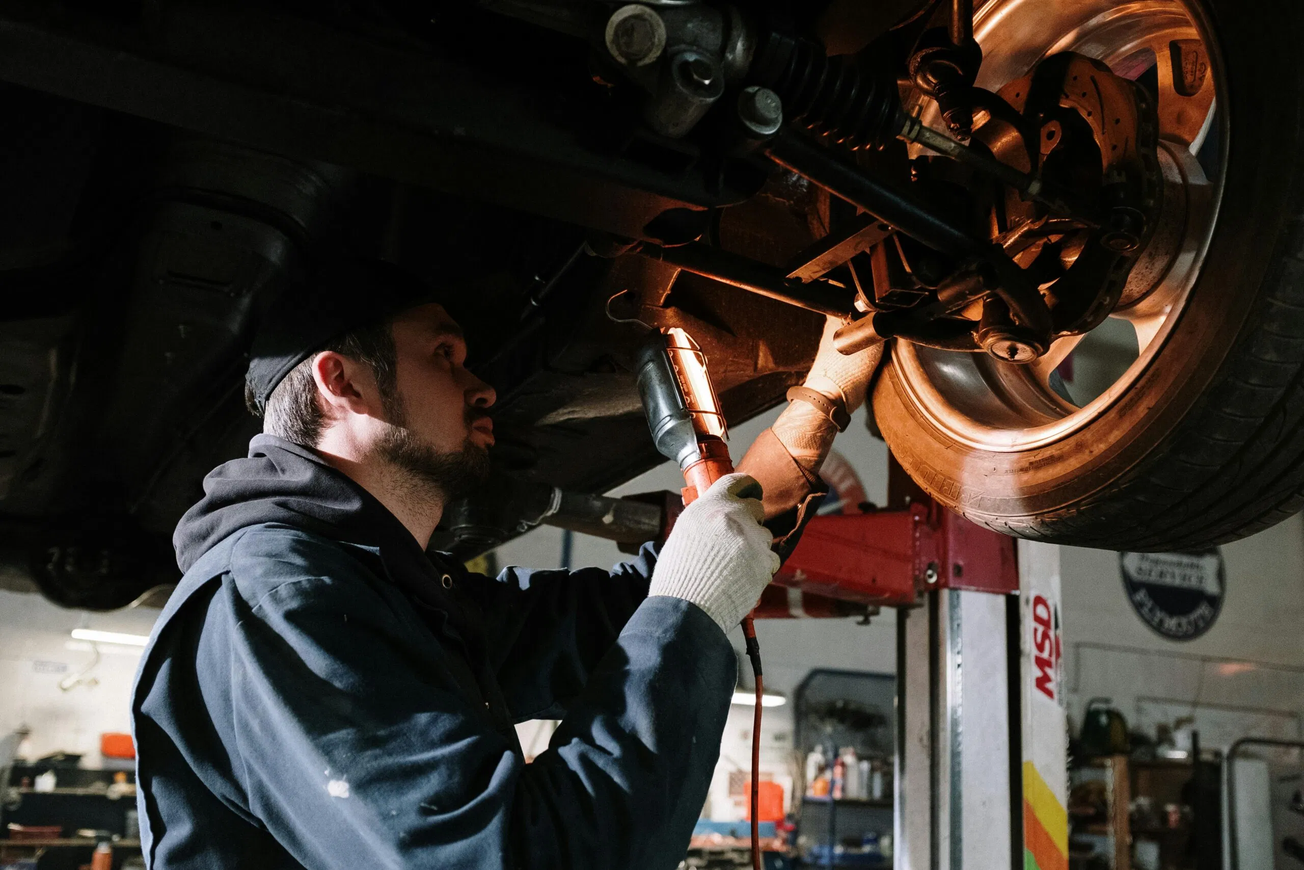 A mechanic inspects and services the undercarriage and brake components of a vehicle using a work light.
