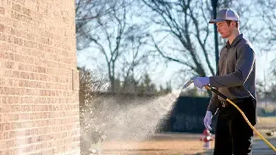 A technician in uniform sprays pest control treatment along the exterior foundation of a brick home, wearing gloves and safety gear to ensure safe and effective perimeter protection against insects and rodents.