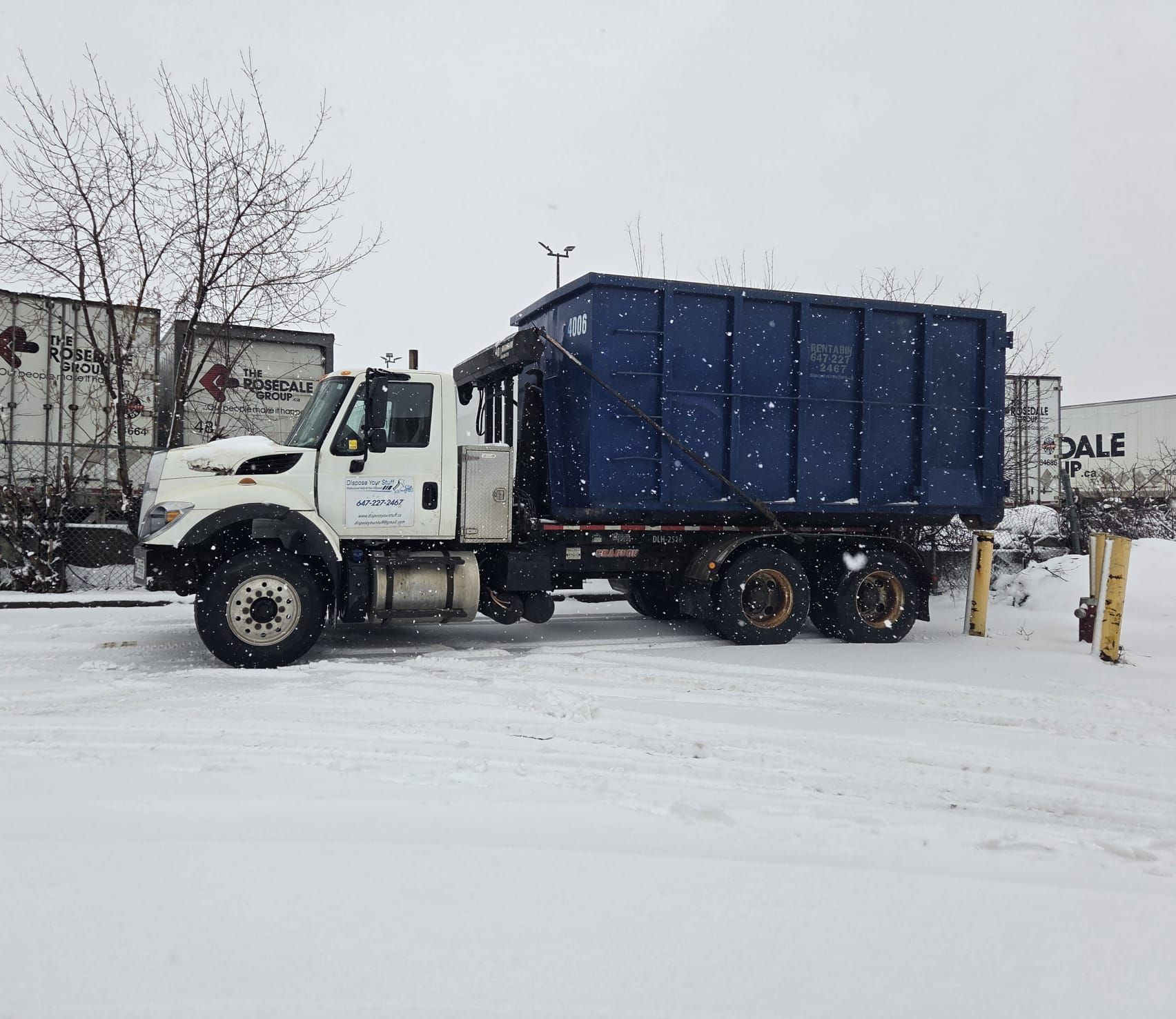 Driveway Safe Dumpsters: Protecting Your Property A Bin4U.ca roll-off truck delivers a large, blue dumpster bin to a commercial site on a snowy day, showcasing the rubber wheel system.