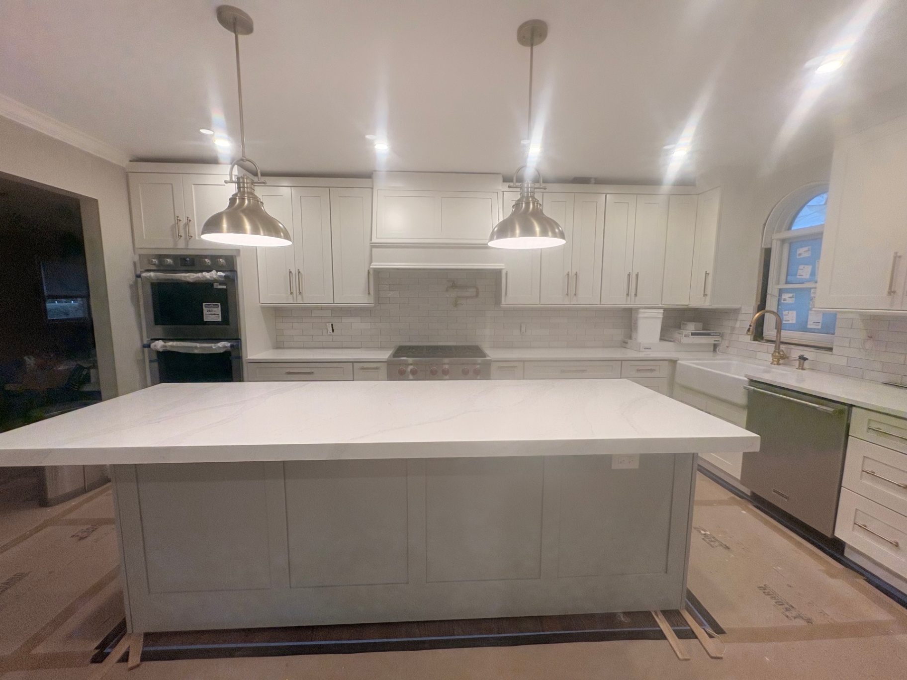 Custom Residential Kitchen by Ragonesi Design LLC This kitchen features shaker-style white cabinetry, a large central island with a quartz countertop, subway tile backsplash, double wall oven, stainless steel fixtures, and a farmhouse sink. Integrated cabinet lighting and pendant lights highlight functional workspaces, while flooring is protected during installation. The design illustrates Ragonesi Design LLC's turnkey approach, blending personalized storage with modern finishes.