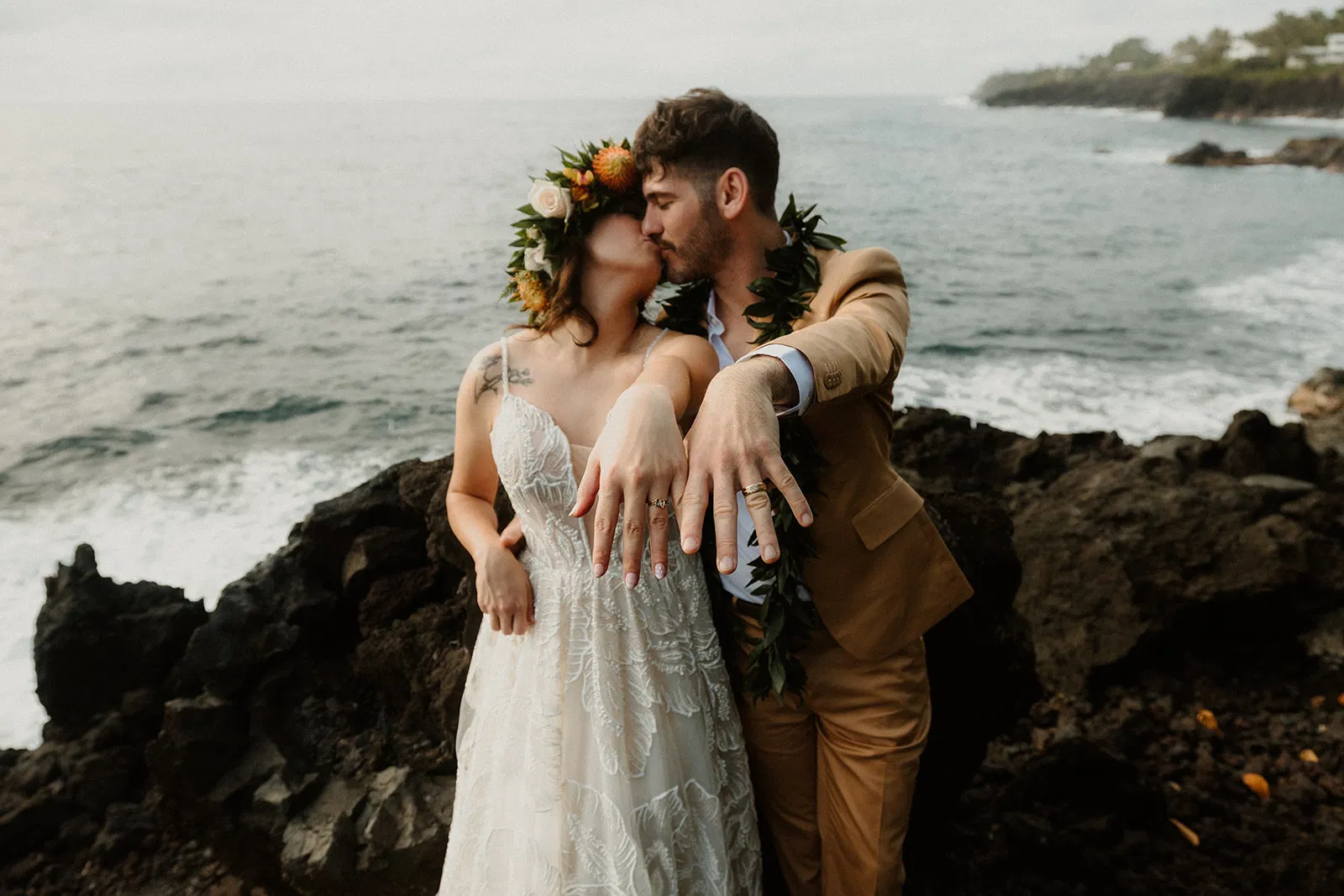 A newlywed couple shares a kiss on the rocky Hawaiian coast, showing off their rings.