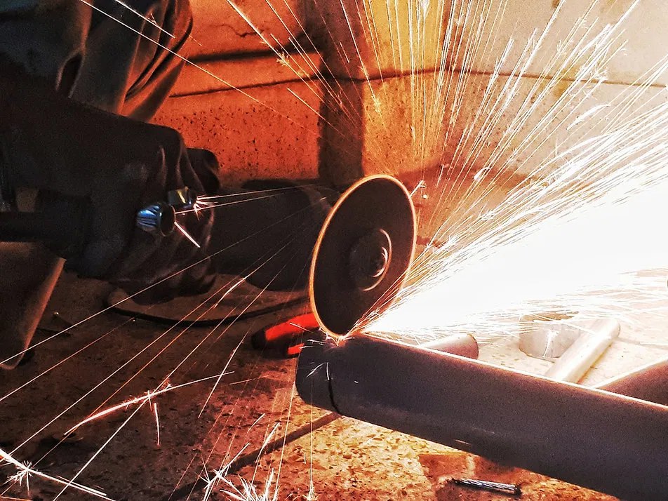 A skilled worker uses a handheld angle grinder to cut through metal piping, producing a shower of sparks, highlighting the technical skill required in the heavy industrial sector.
