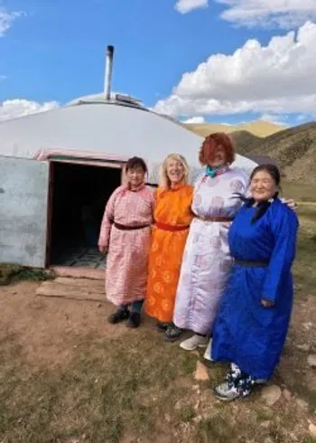Four women in traditional Mongolian dress stand arm-in-arm in front of a ger, smiling under an open sky, symbolizing global sisterhood.
