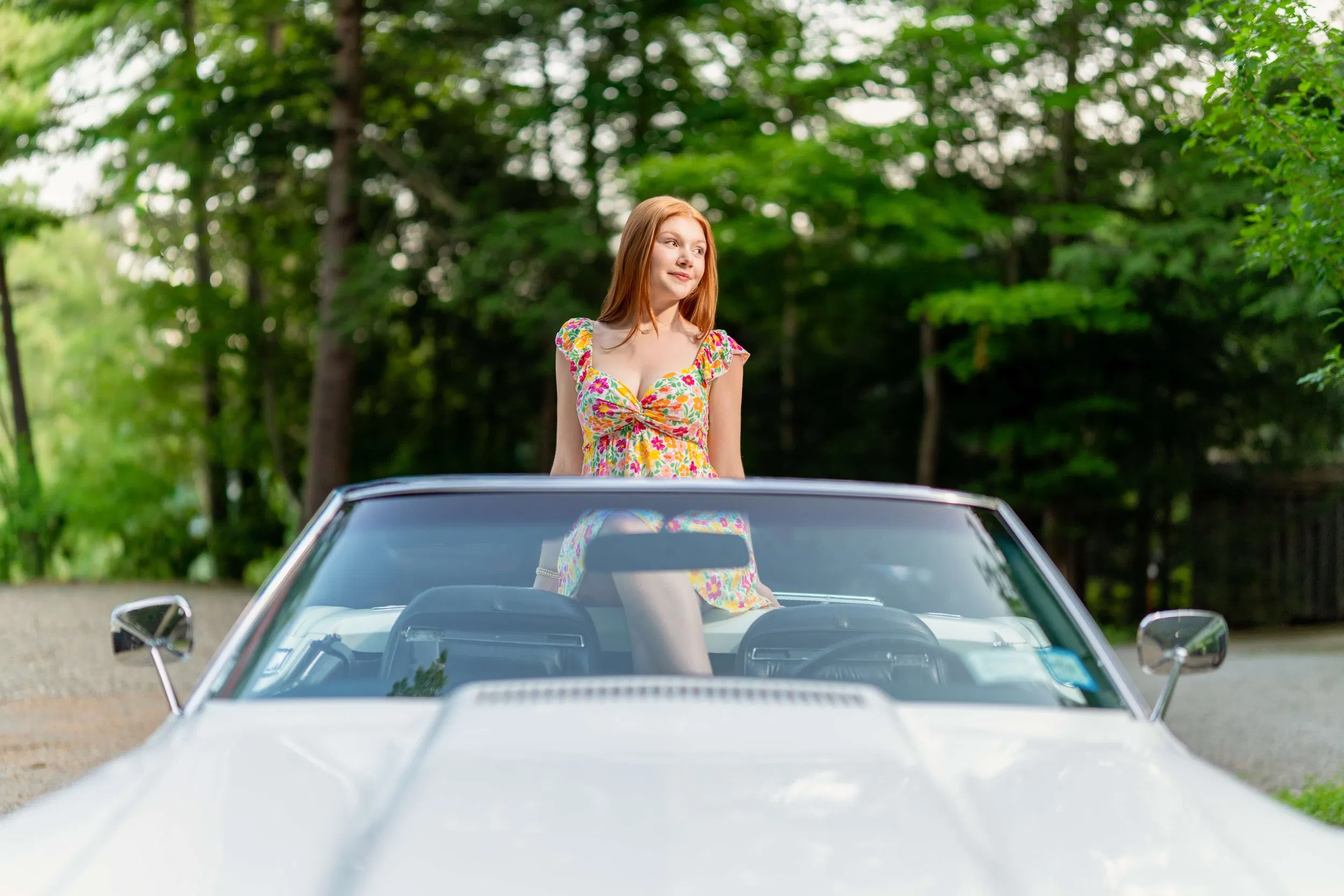 A confident young woman in a colorful, floral dress is sitting on the back of a white convertible car, looking off to the side with a relaxed, radiant expression.