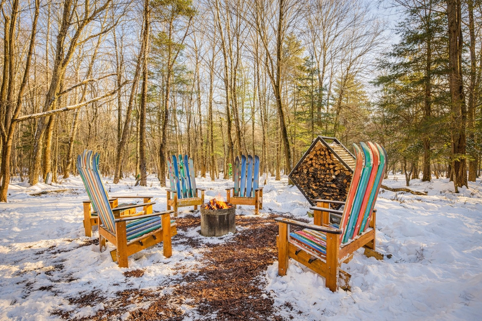 [HERO] The real ski-chair fire pit at Bearly Roughing It — four Adirondack chairs made from vintage skis around the fire pit in the snow with the firewood rack