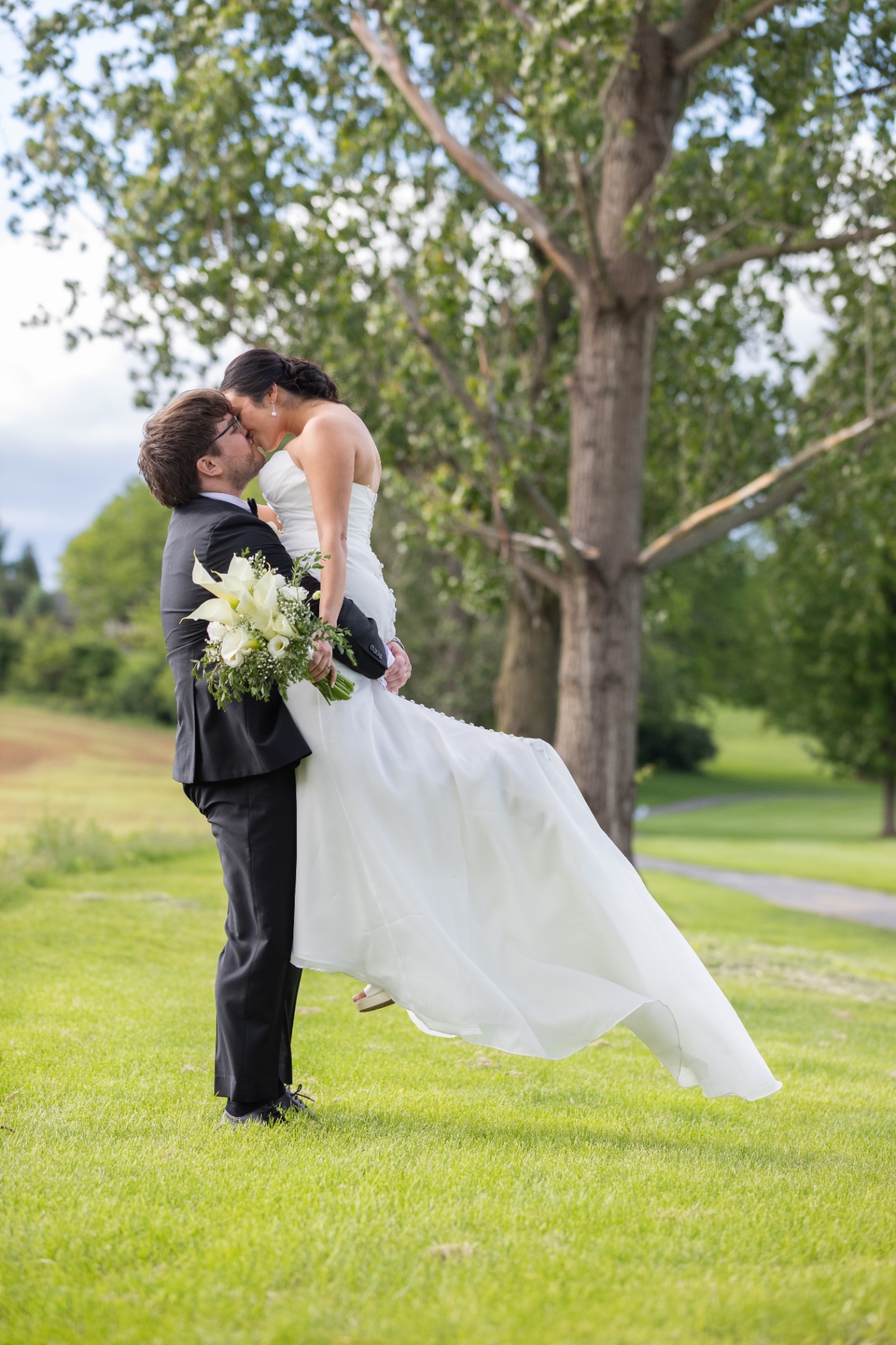 A groom in a black suit lifts the bride as they kiss on a bright outdoor lawn