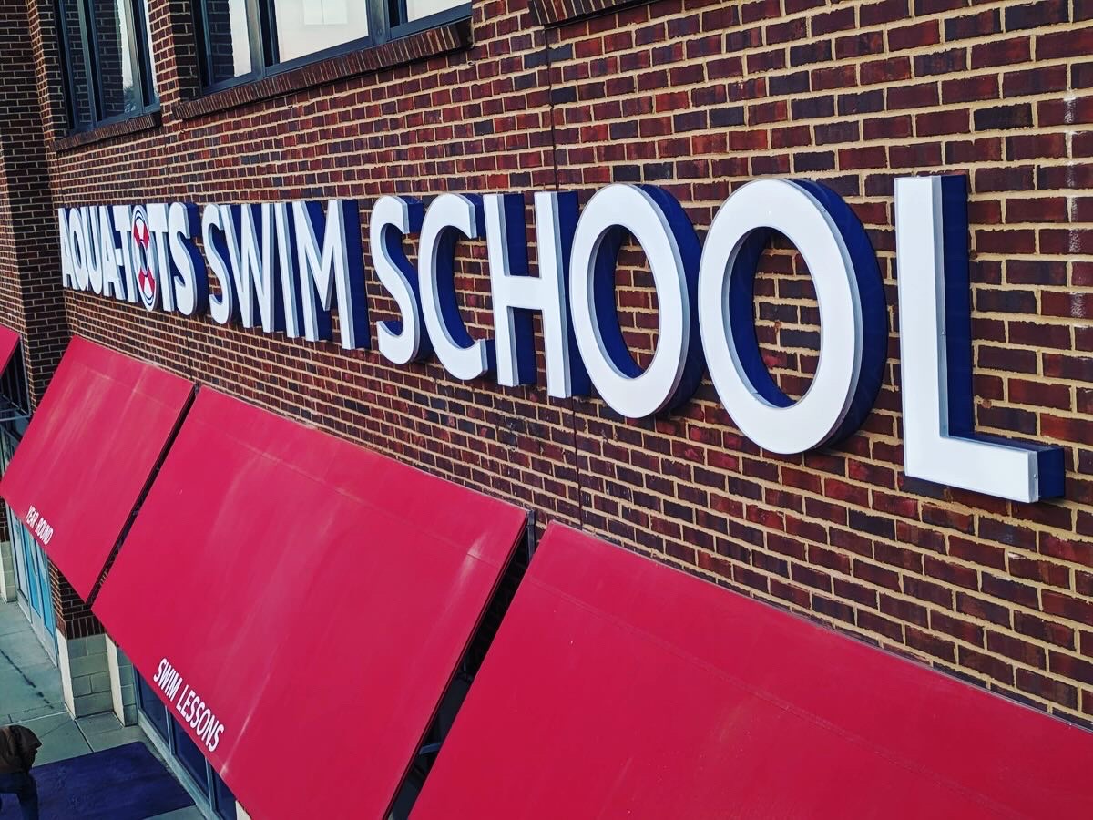 AQUA-TOTS SWIM SCHOOL Channel Letter Sign Custom lighted channel letter sign installed on a brick exterior, featuring bold, three-dimensional white and blue letters for 