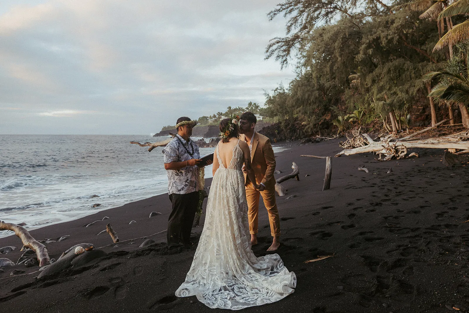 A barefoot bride and groom exchange vows on a dramatic black sand beach at sunset, surrounded by lush greenery and ocean waves.