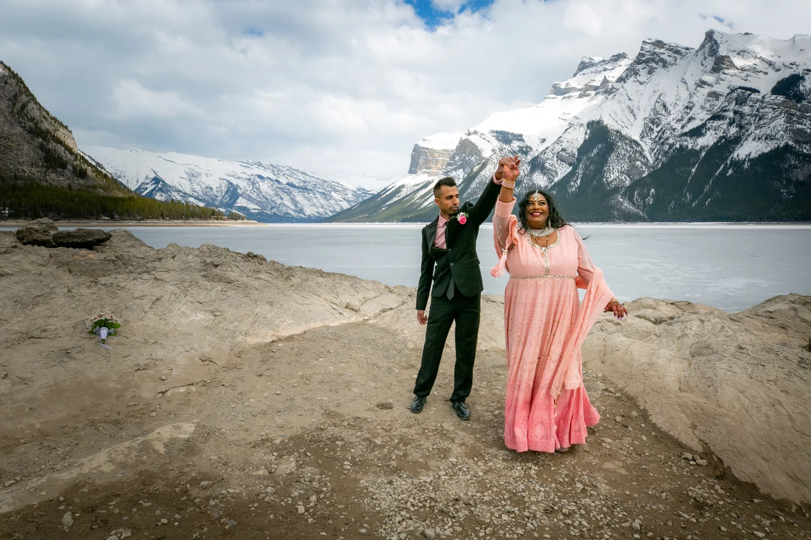 Elopement at Lake Minnewanka A couple celebrates their elopement by Lake Minnewanka, with snow-capped Rocky Mountains in the background. The bride wears a pink dress, and the groom is in a dark suit with a rose boutonniere. A bridal bouquet rests on the rocks beside the lake.