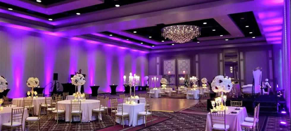 Elegant Ballroom Event by Modish Entertainment Elegant ballroom setup with purple uplighting, round tables adorned with floral centerpieces, gold chiavari chairs, a large chandelier, and a clear dance floor. Professional event lighting, decor, and arrangement showcase Modish Entertainment's upscale event planning capabilities for weddings and special occasions.