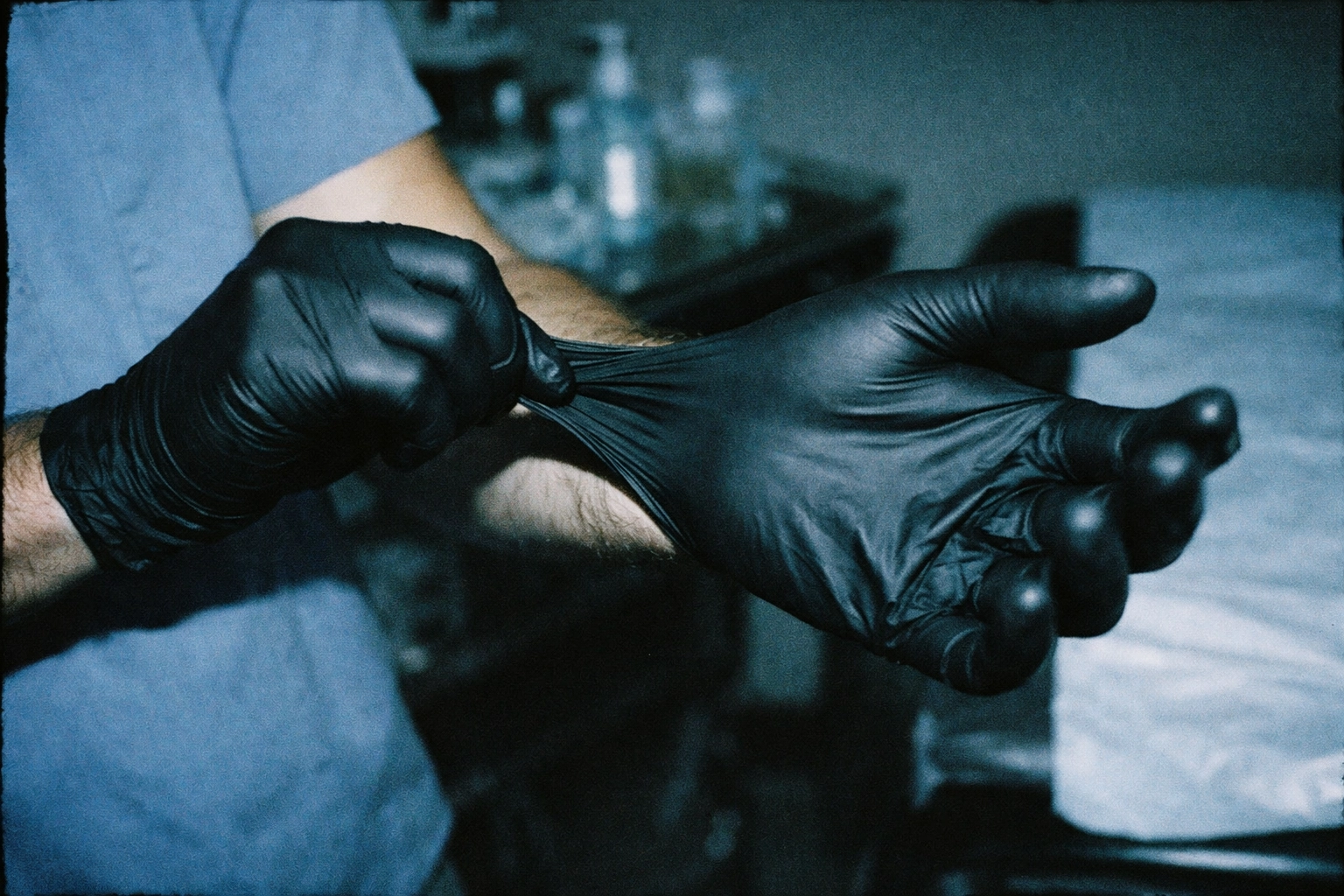A black nitrile glove being snapped onto a wrist in a hospital exam room—professional, controlled, slightly eerie.