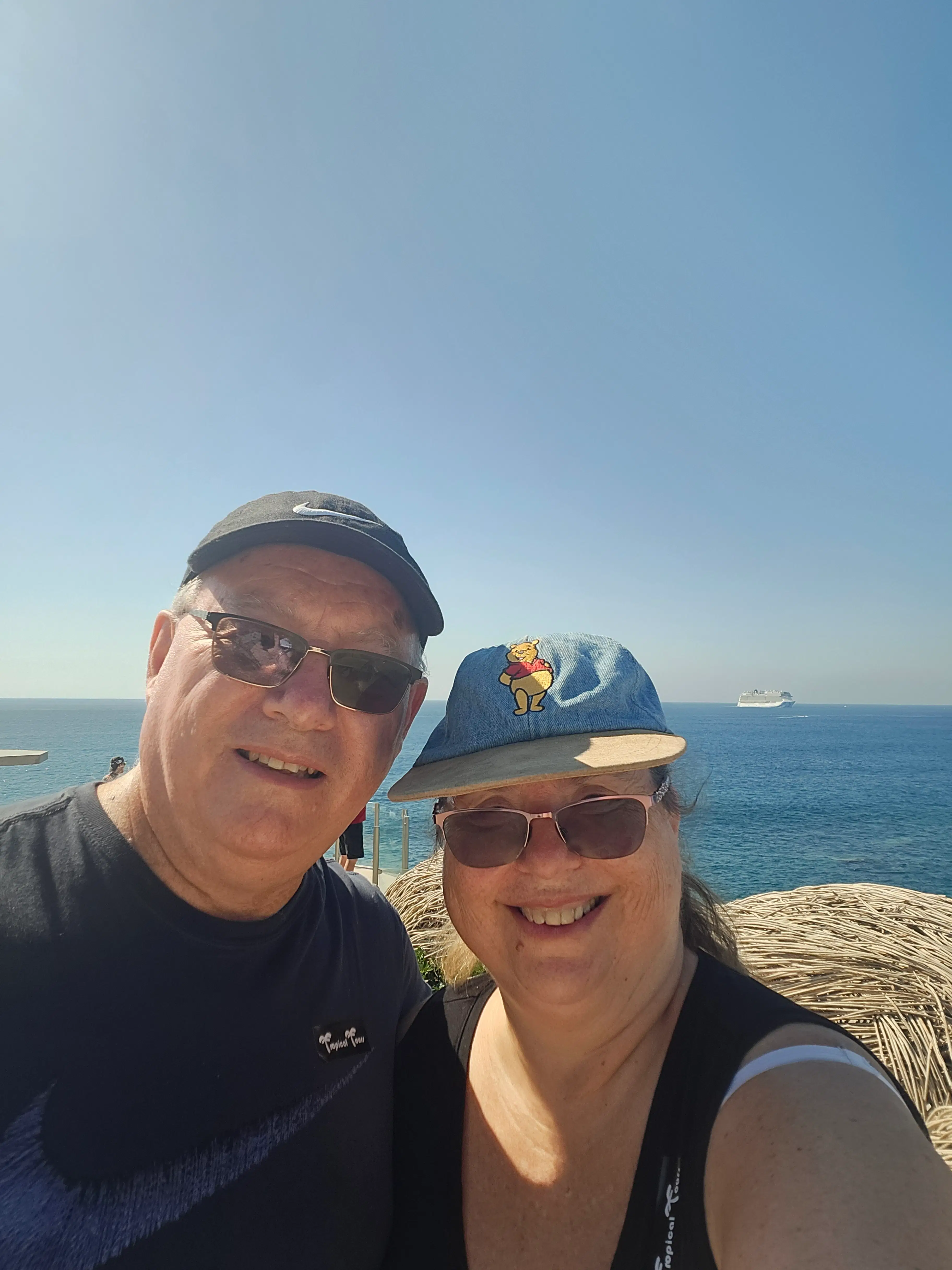 A smiling couple taking a selfie with a cruise ship in the background