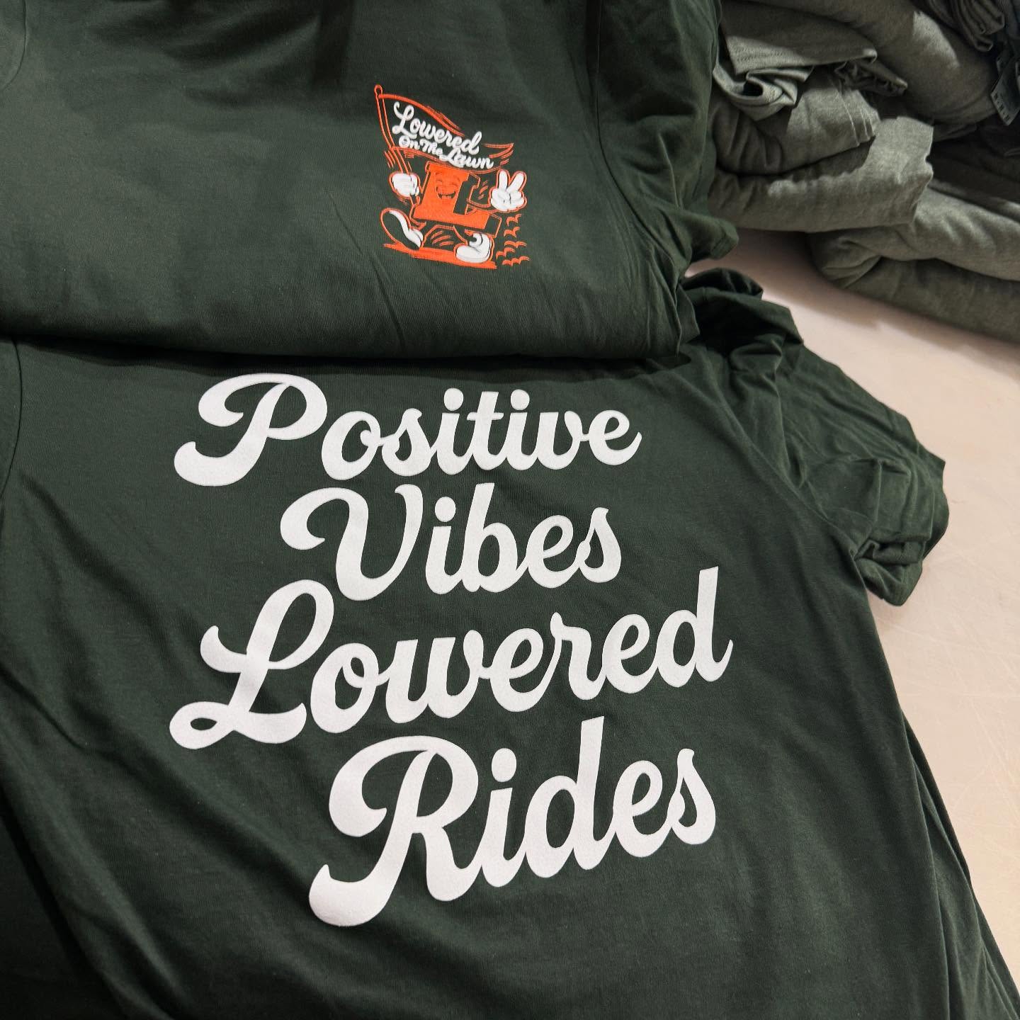 Dark Green Custom Lowered Rides T-Shirts Dark green custom-printed t-shirts featuring bold white script 'Positive Vibes Lowered Rides' on the front and a small graphic with 'Lowered on the Farm' on the upper chest, produced in-house and folded on a work surface ready for pickup or shipping.