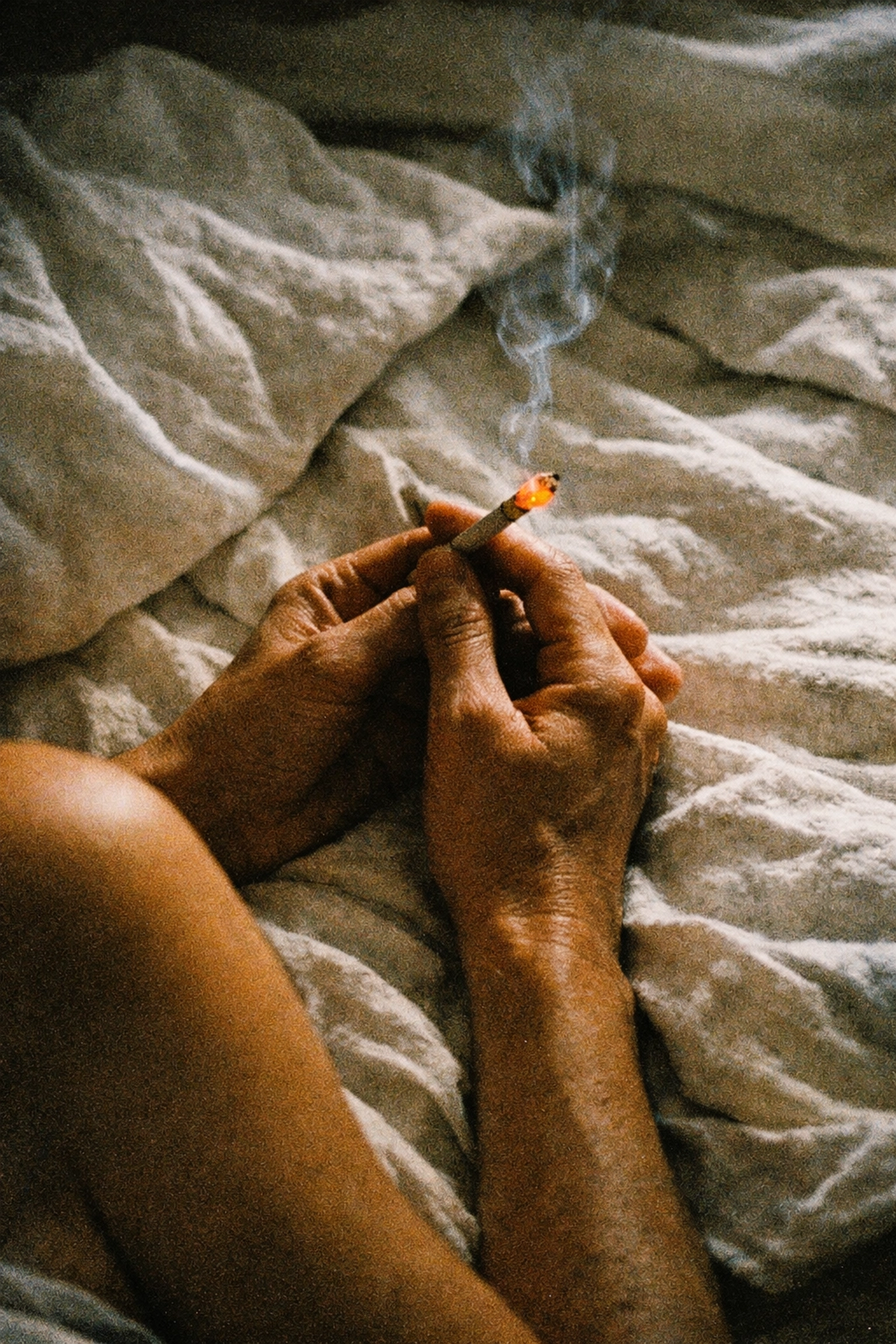 Close-up of hands holding lit cigarette on bed showing the aesthetic ritual of smoking