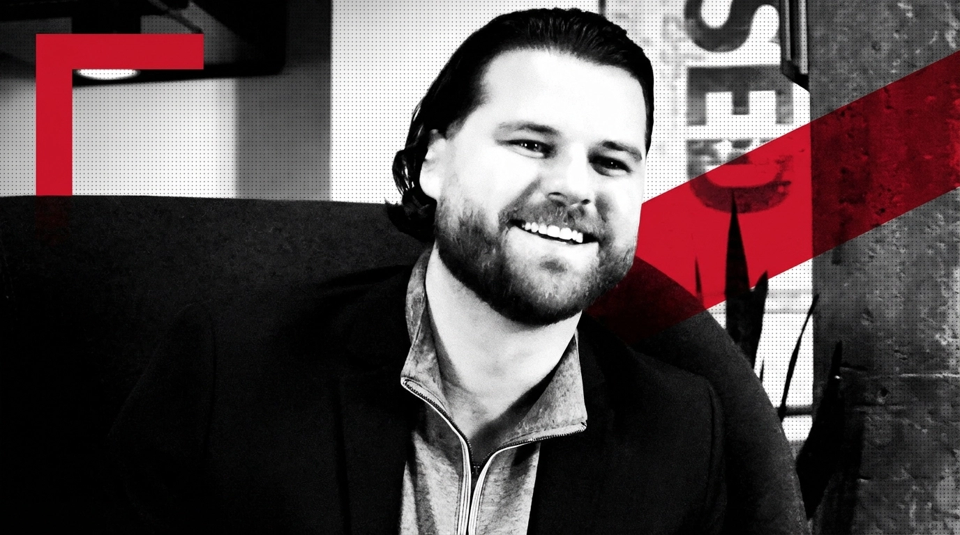 Smiling Man in Modern Office A smiling man in business casual attire sits on a red chair in a modern office space, reflecting a welcoming and professional environment tailored to support real estate agents in building their businesses. A partial motivational sign is visible in the background.