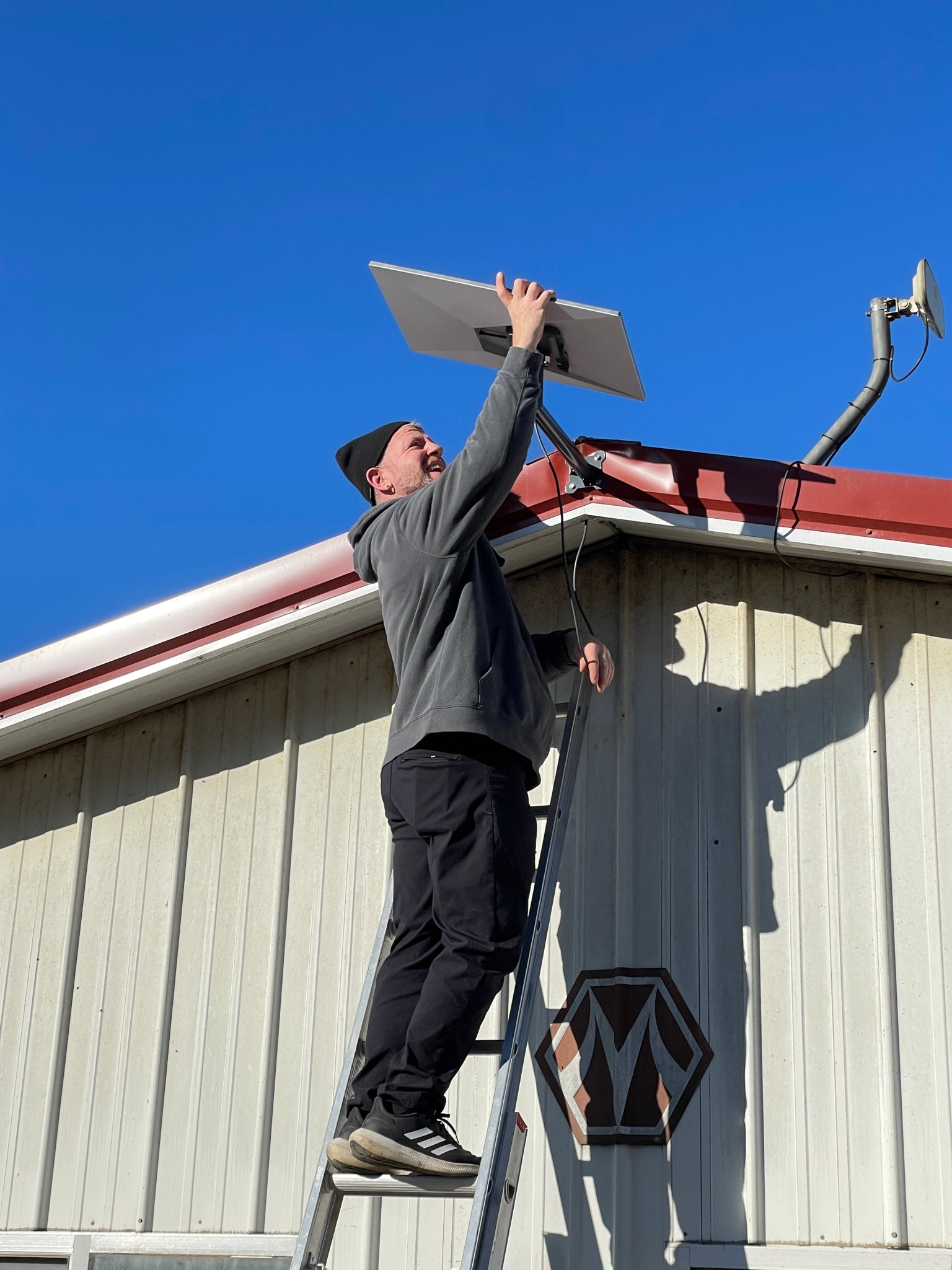 Technician on a ladder installing a Starlink dish on a metal farm building in Michigan, showing professional equipment and careful mounting work.
