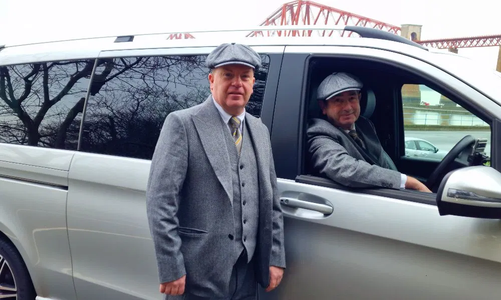 Two professional chauffeurs dressed in grey suits and flat caps standing by a silver executive vehicle with the Forth Bridge in the background