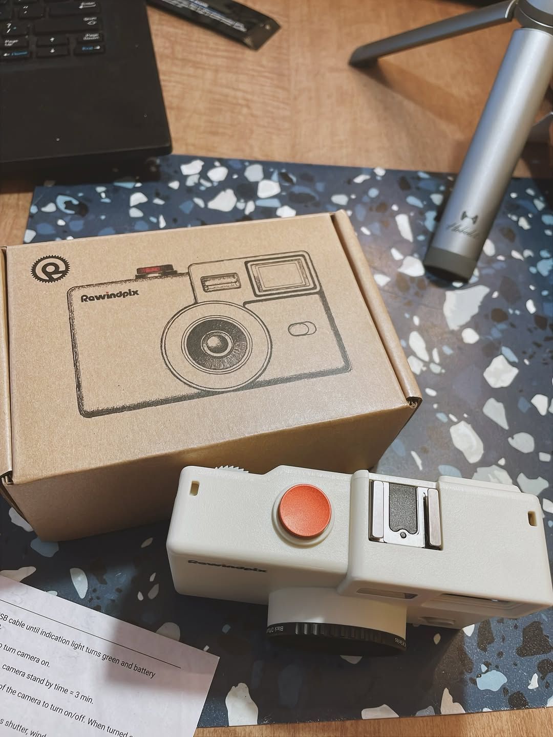 A compact point-and-shoot Rewindpix camera named AJ1ZDPa05kb features a large red shutter button. The camera is displayed outside its illustrated box, resting on a speckled mat beside printed instructions, a laptop, and a silver tripod. The scene highlights new gear arrivals for photography enthusiasts.