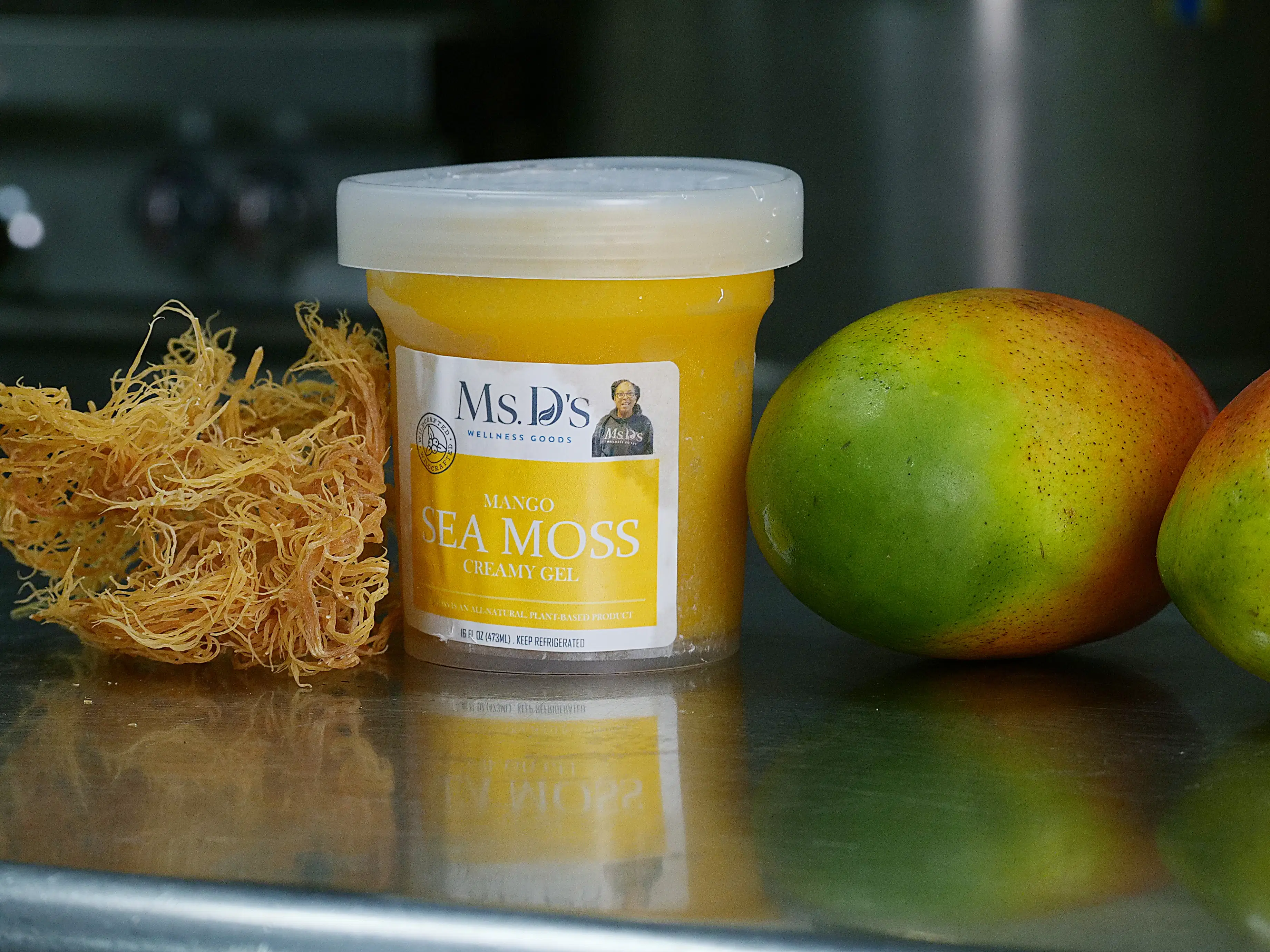 Product photo of Ms. D’s Mango Sea Moss Creamy Gel in a 16oz jar, featured alongside fresh raw gold sea moss and whole ripe mangoes on a stainless steel prep counter, highlighting the real, premium-quality ingredients used in the naturally flavored wellness gel.