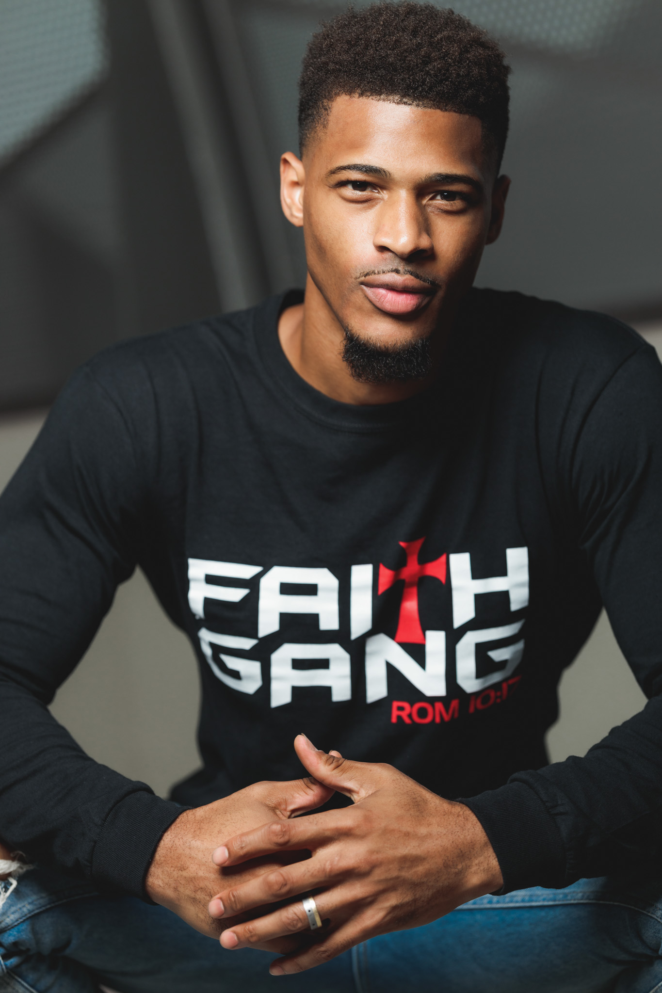 Faith Gang Sweatshirt Model A male model wears a black long-sleeve sweatshirt featuring bold white 'FAITH GANG' lettering with a red cross and 'ROM 10:17' scripture reference, styled with blue jeans. The garment highlights TruVine Apparel's signature Christian streetwear aesthetic and premium quality.