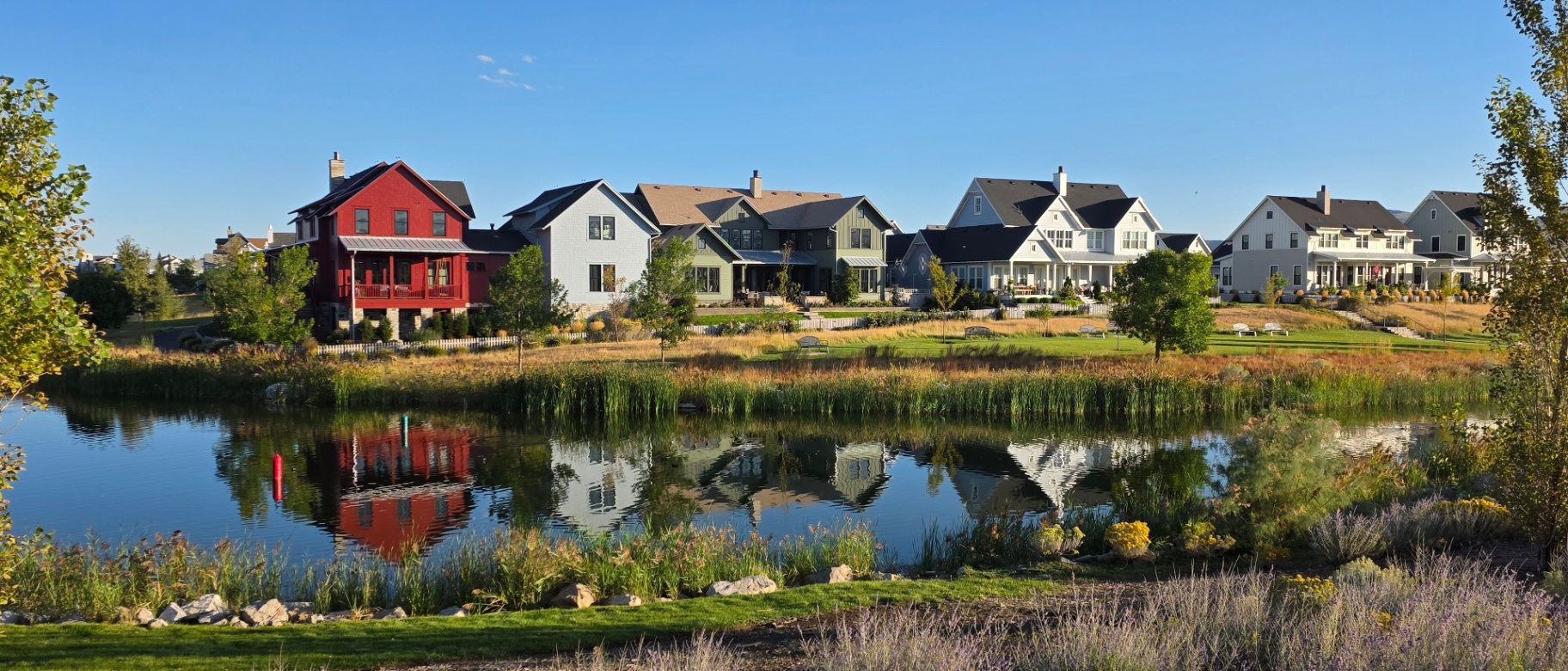 A row of modern, single-family homes in a suburban Utah neighborhood, each featuring landscaped yards, front porches, and a scenic pond with reflections in the foreground.