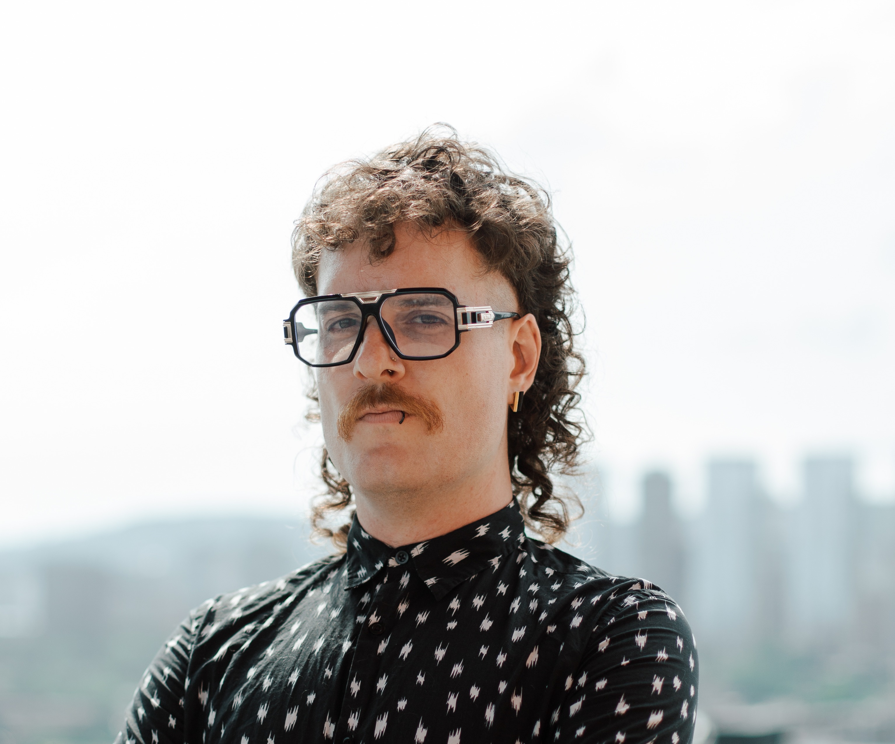 Tyler St Germain A confident SEO pro stands outdoors with a city skyline in the background, wearing large glasses and a patterned shirt. This image can represent the creative and approachable SEO team behind Twenty West Media's web design and marketing solutions.