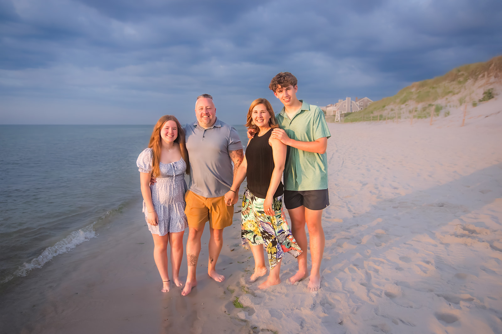 A family of four stands barefoot on a Cape Cod beach at sunset, posing together with relaxed smiles.