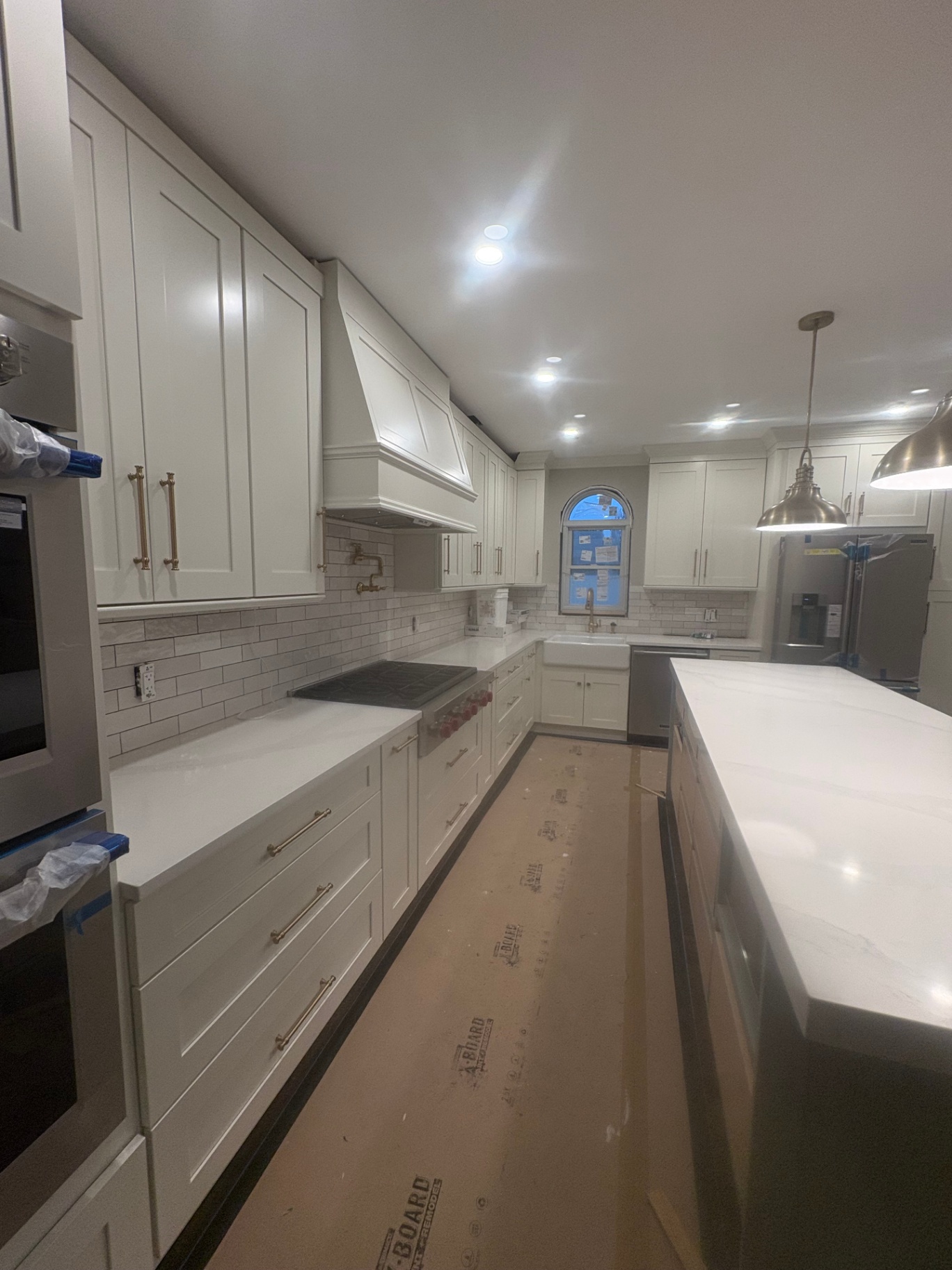 Custom Kitchen Custom kitchen featuring full-height white shaker cabinets, quartz countertops, subway tile backsplash, integrated hood, gold hardware, chef's range, farmhouse sink, and large center island. LED recessed lighting and pendant fixtures provide layered illumination; millwork and cabinetry are crafted to client specifications in our in-house shop.