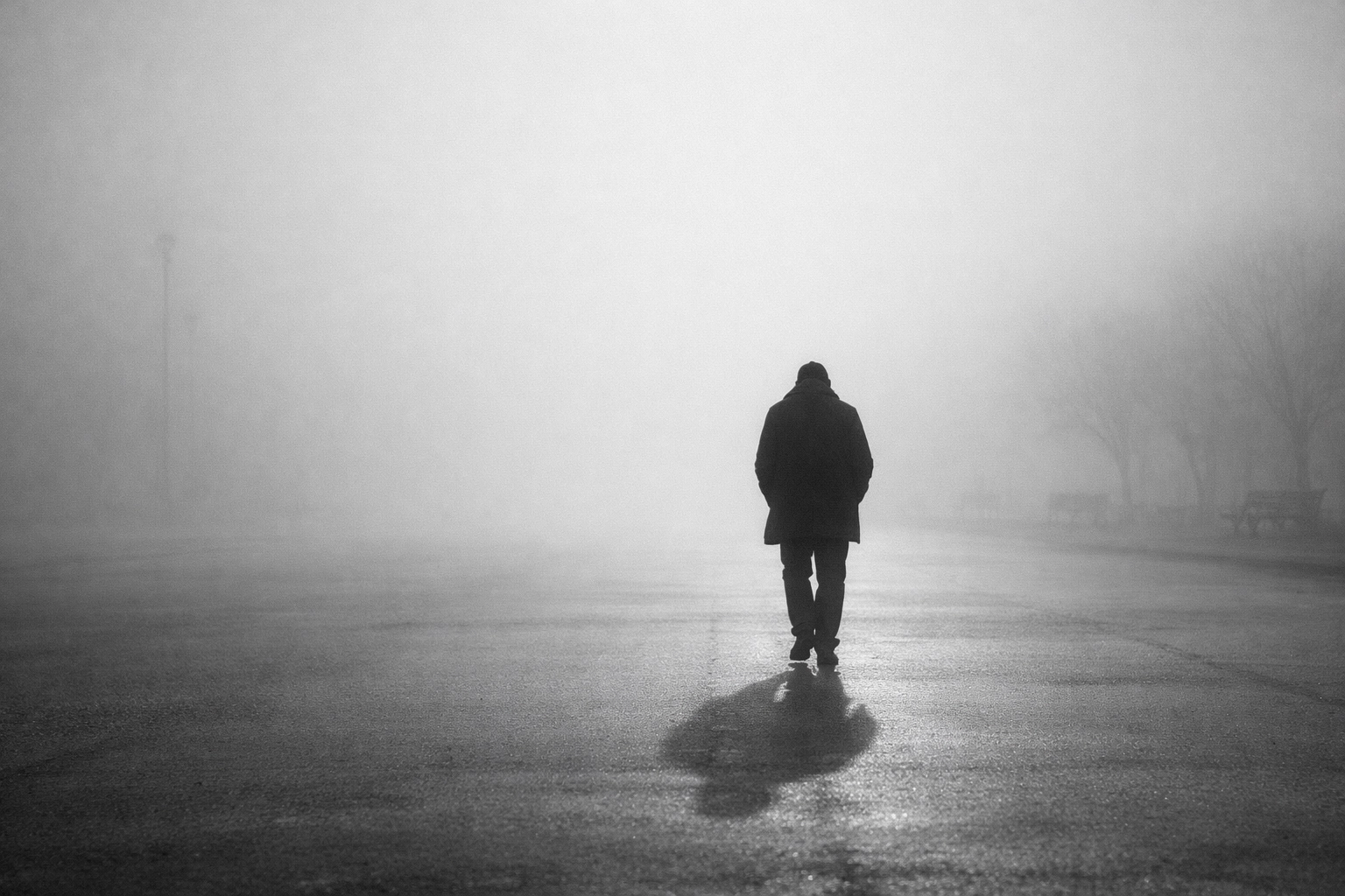 Black and white wide shot of a lone adult walking through a quiet, misty empty street, capturing the feeling of going through the motions and wading through treacle.