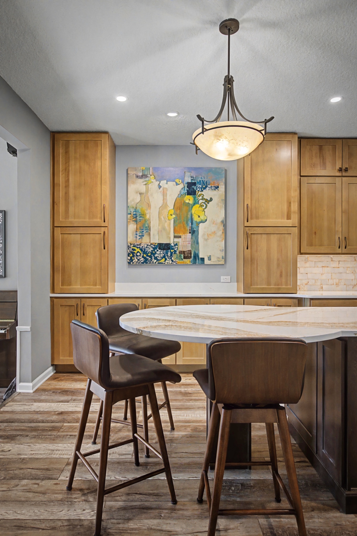 A modern kitchen featuring natural wood cabinetry, a large stone-topped island with seating, pendant lighting, and contemporary art.