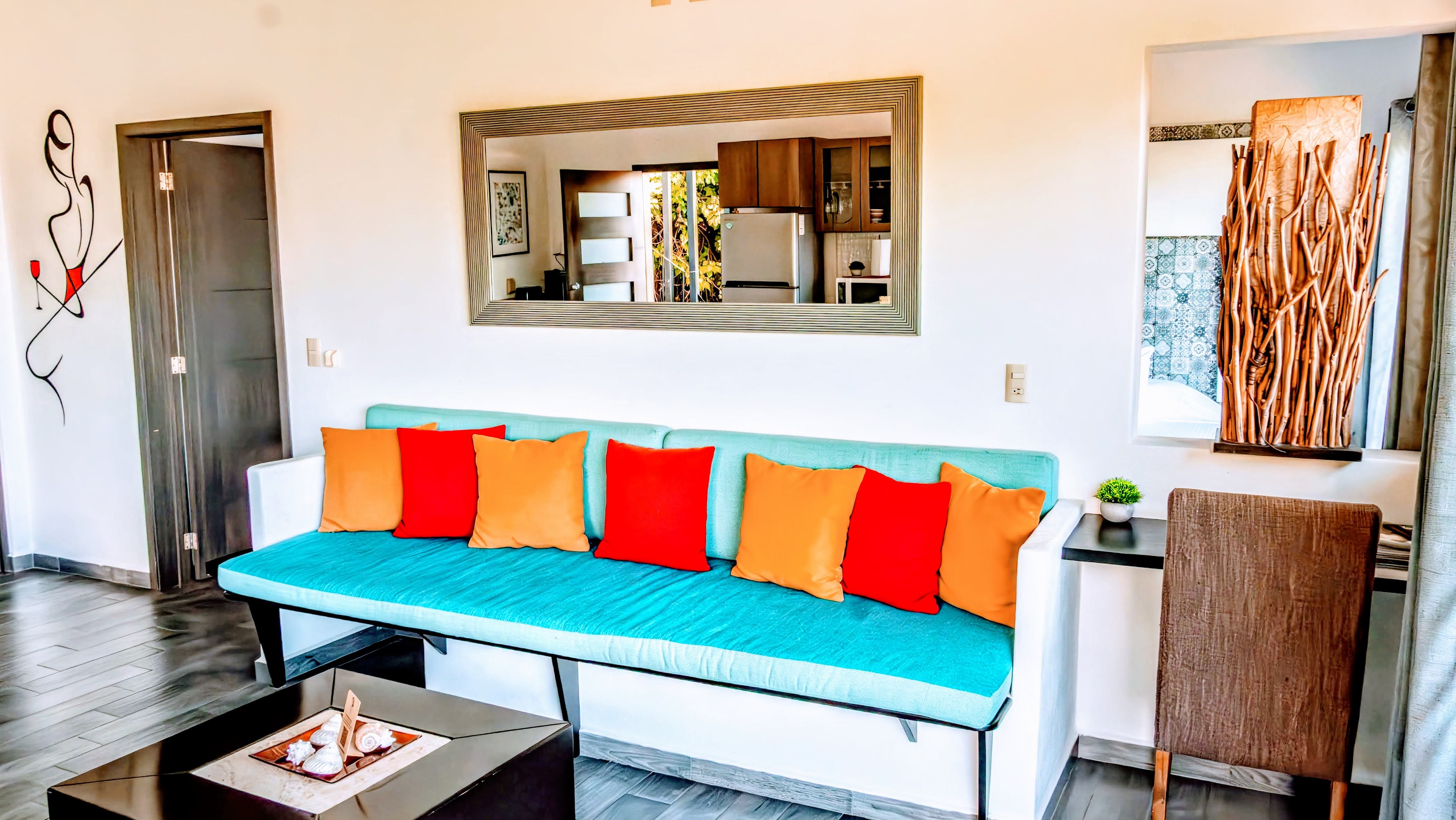 Bright, modern living room in a Puerto Vallarta condo A living room featuring a turquoise sofa with vibrant orange and red pillows, a large wall mirror reflecting the open kitchen, artistic wall decor, natural wood accents, and a cozy workspace that adds local character and comfort.