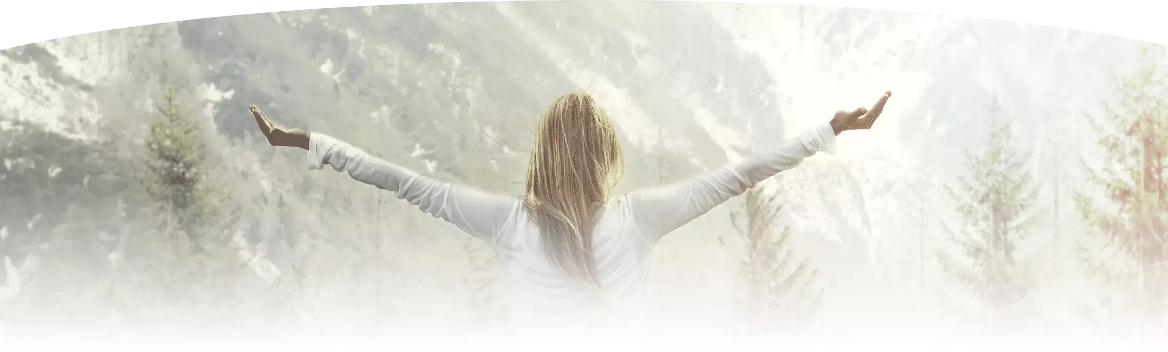 A woman stands outdoors facing a serene mountain landscape, arms outstretched in a gesture of openness and vitality.