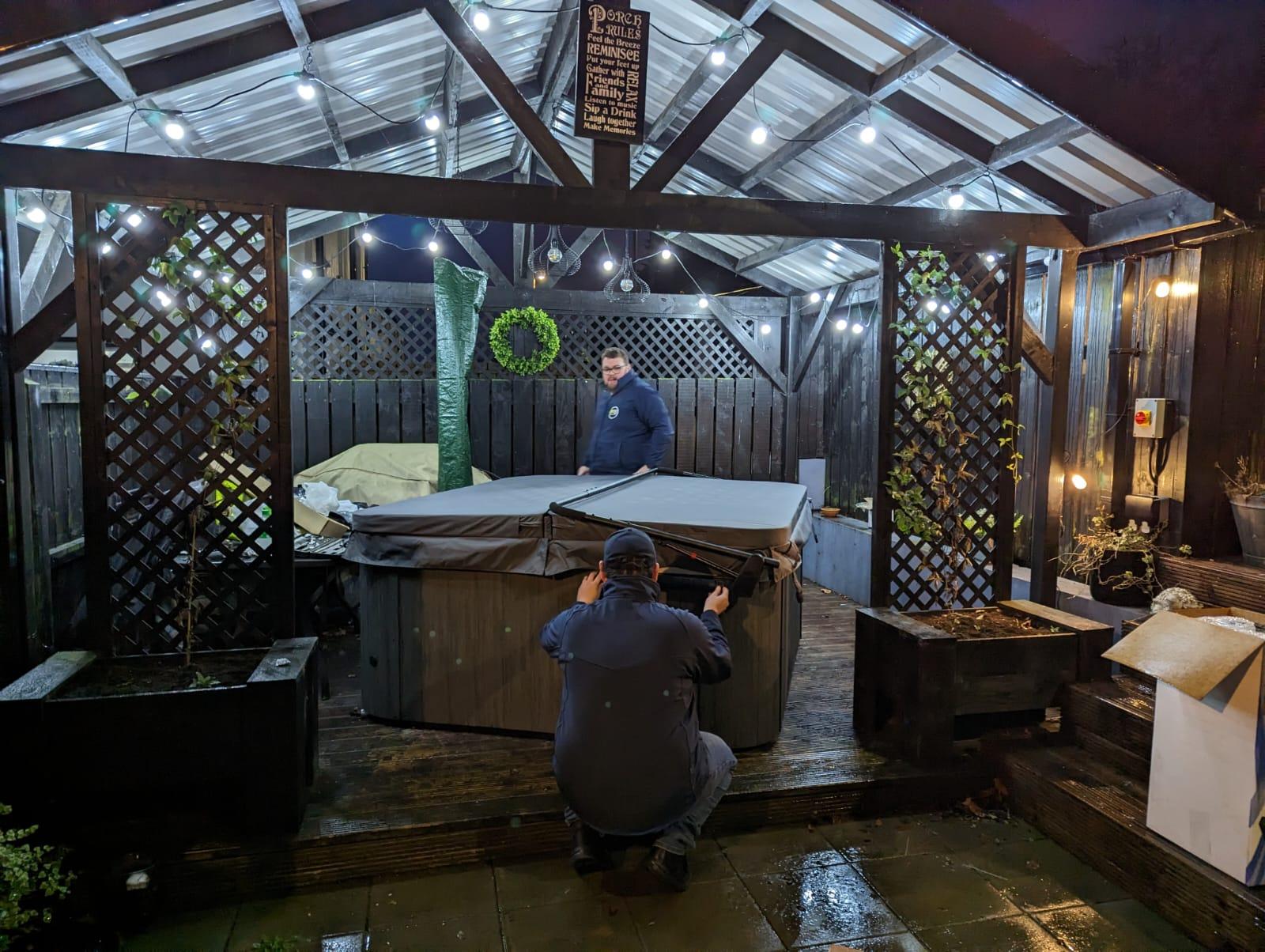 Fiesta Spas technicians installing an Artesian Spa hot tub with an insulated cover under a lit outdoor pergola