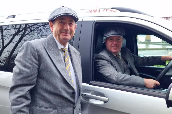 Two Aura Journeys chauffeurs in matching grey suits and flat caps standing by a luxury Mercedes-Benz
