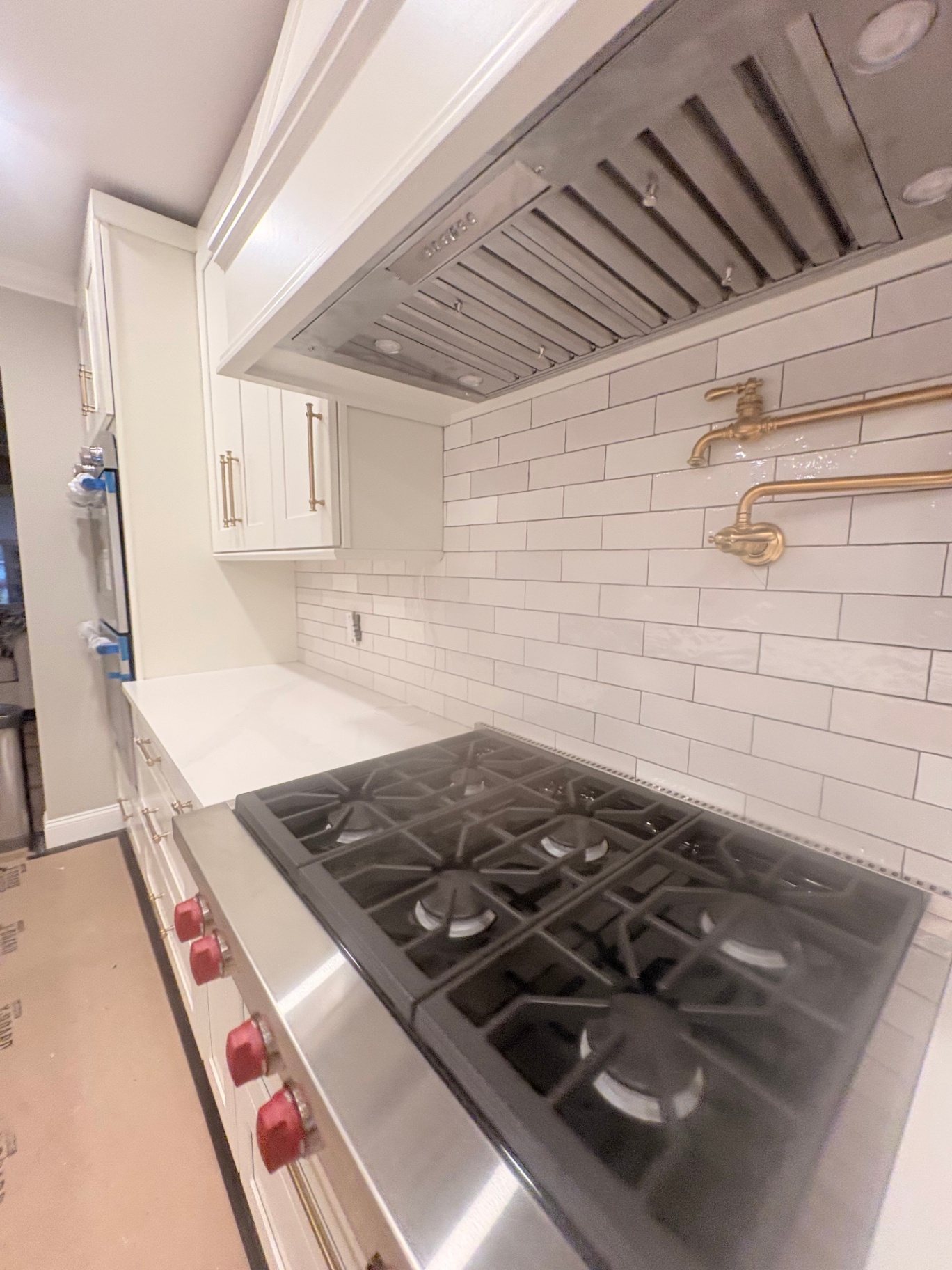 Custom Residential Kitchen Install A kitchen installation featuring a professional-grade 6-burner gas cooktop, white shaker-style cabinets with brass bar pulls, white quartz countertops, subway tile backsplash, contemporary brass pot filler, and a high-powered stainless-steel range hood, all meticulously designed for premium functionality and style.