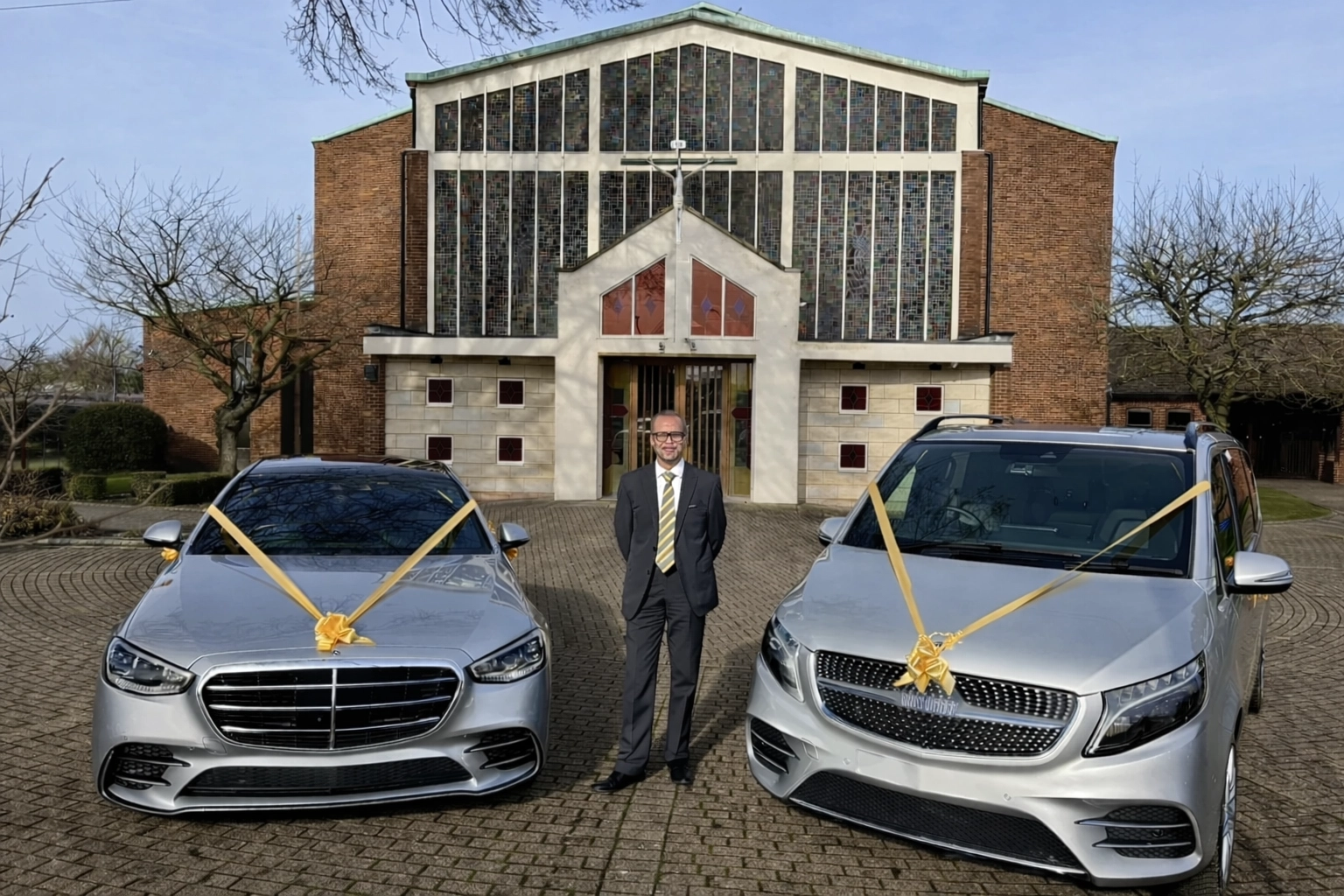 Two silver Mercedes-Benz wedding cars decorated with gold wedding ribbons are parked outside a church entrance. A professionally dressed chauffeur stands between them.